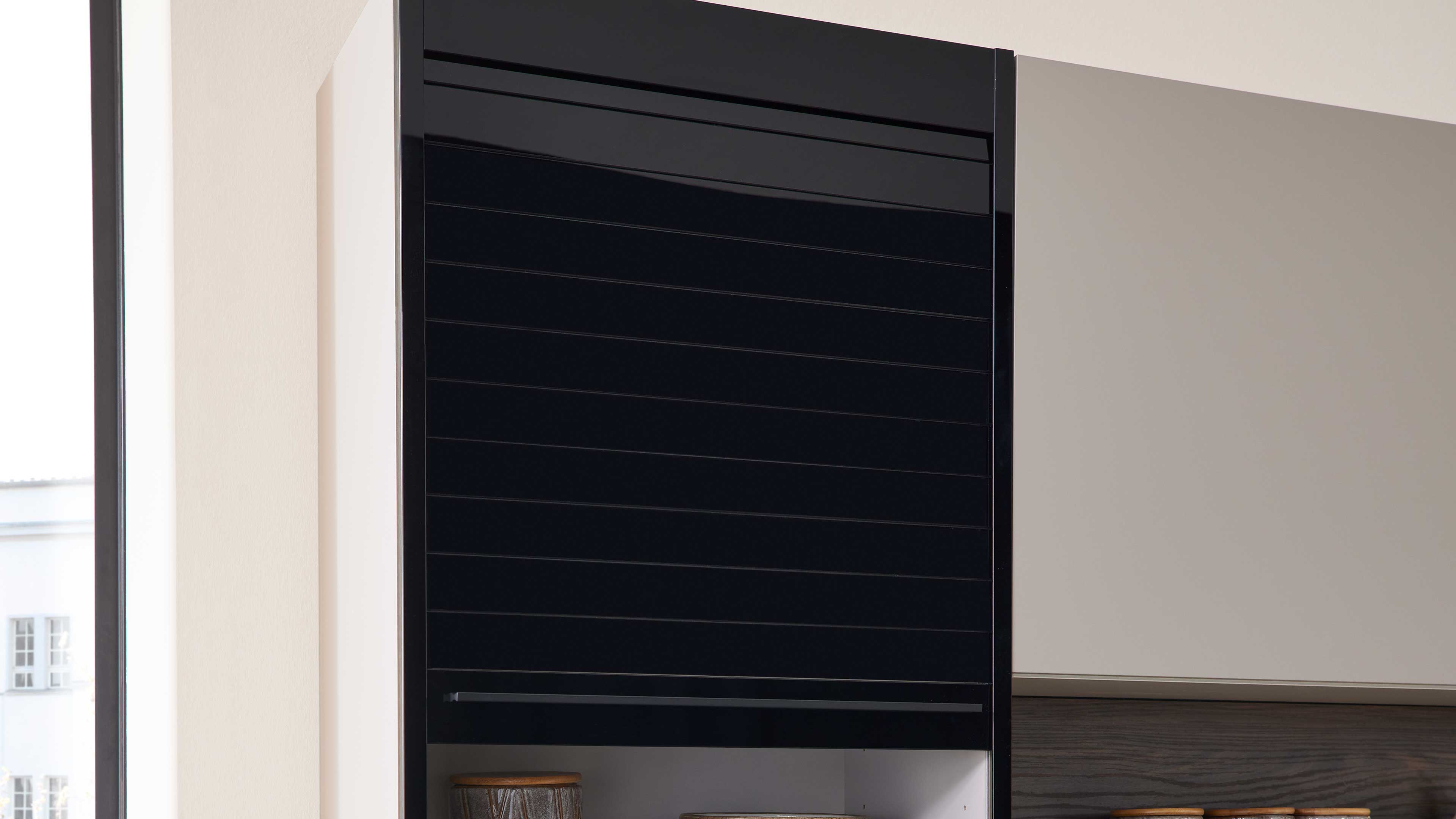 Schwarze hochglänzende Schrankfront mit horizontalen Lamellen und schmalem Griff, daneben helle Front und offenes Fach mit zwei runden Gefäßen.