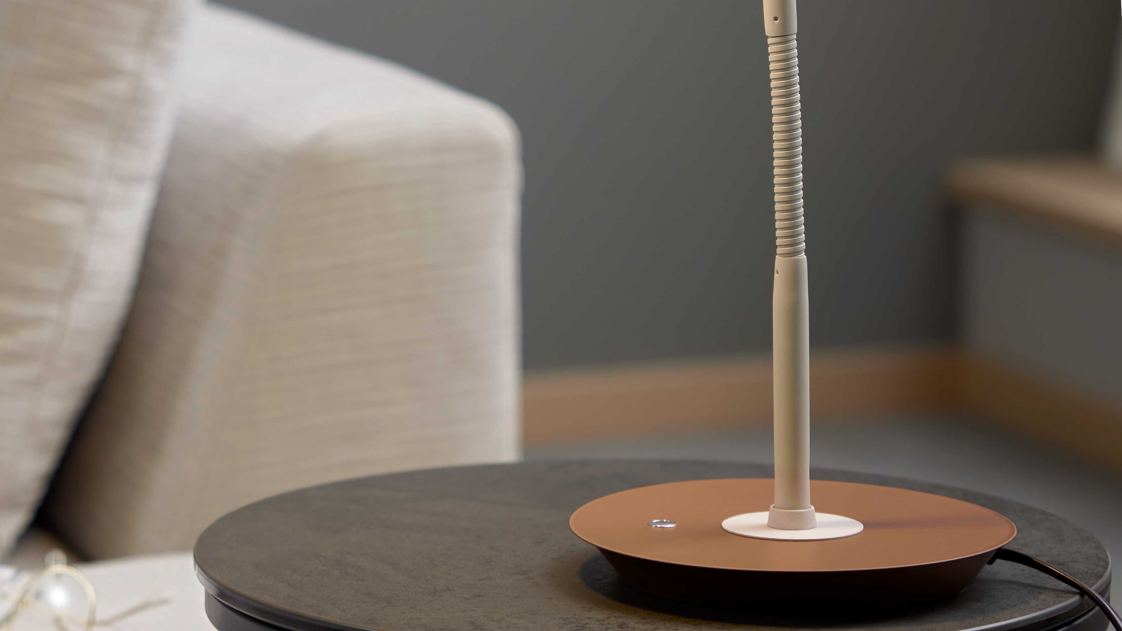 Tischlampe mit rundem, rostbraunem Sockel und flexiblem Schwanenhals auf dunklem, rundem Beistelltisch neben einem hellen Sofa.