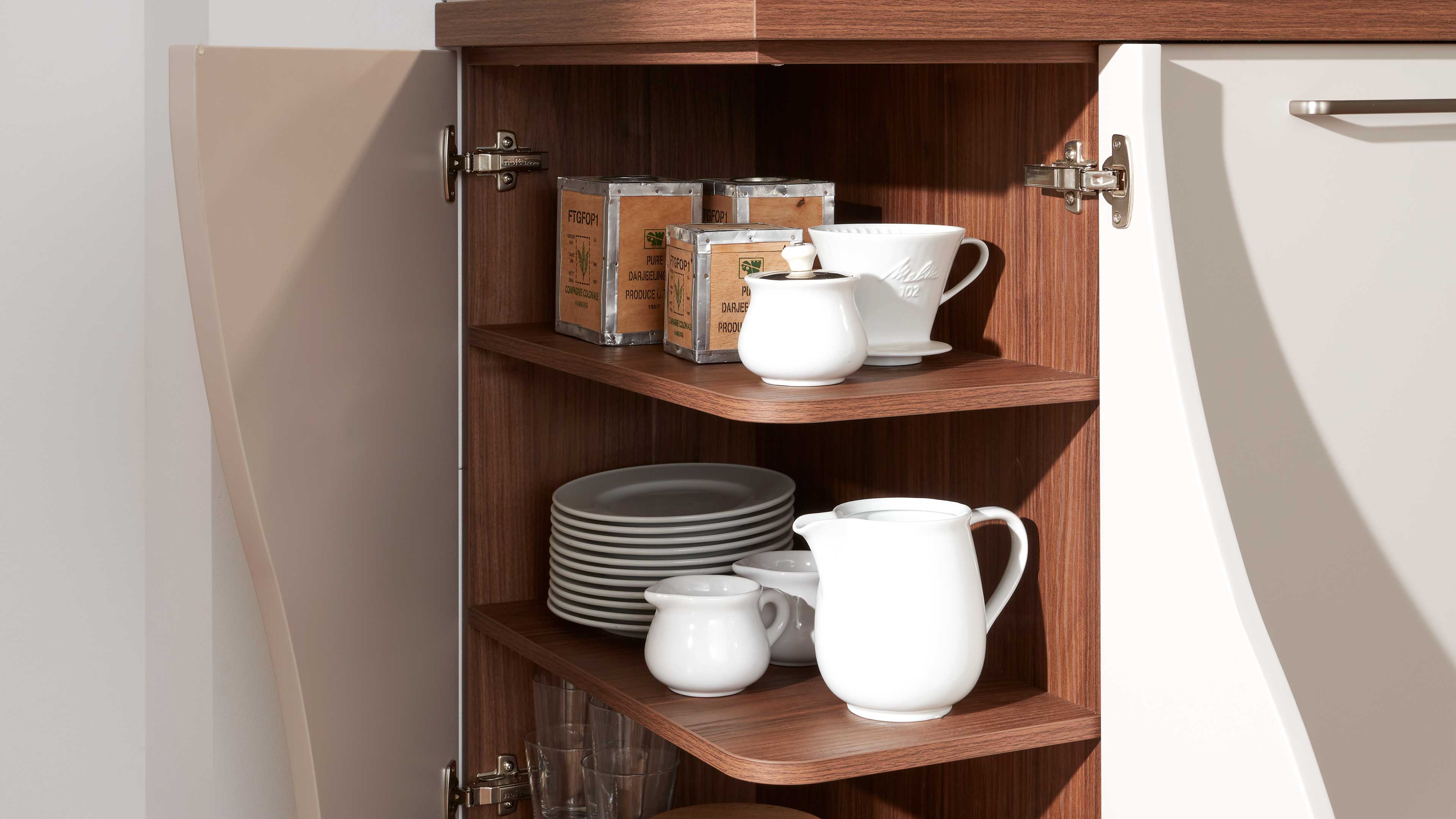 Offenes Küchenschrankfach in Holzoptik mit weißen Porzellangefäßen, gestapelten Tellern, Gläsern und kleinen Teedosen.
