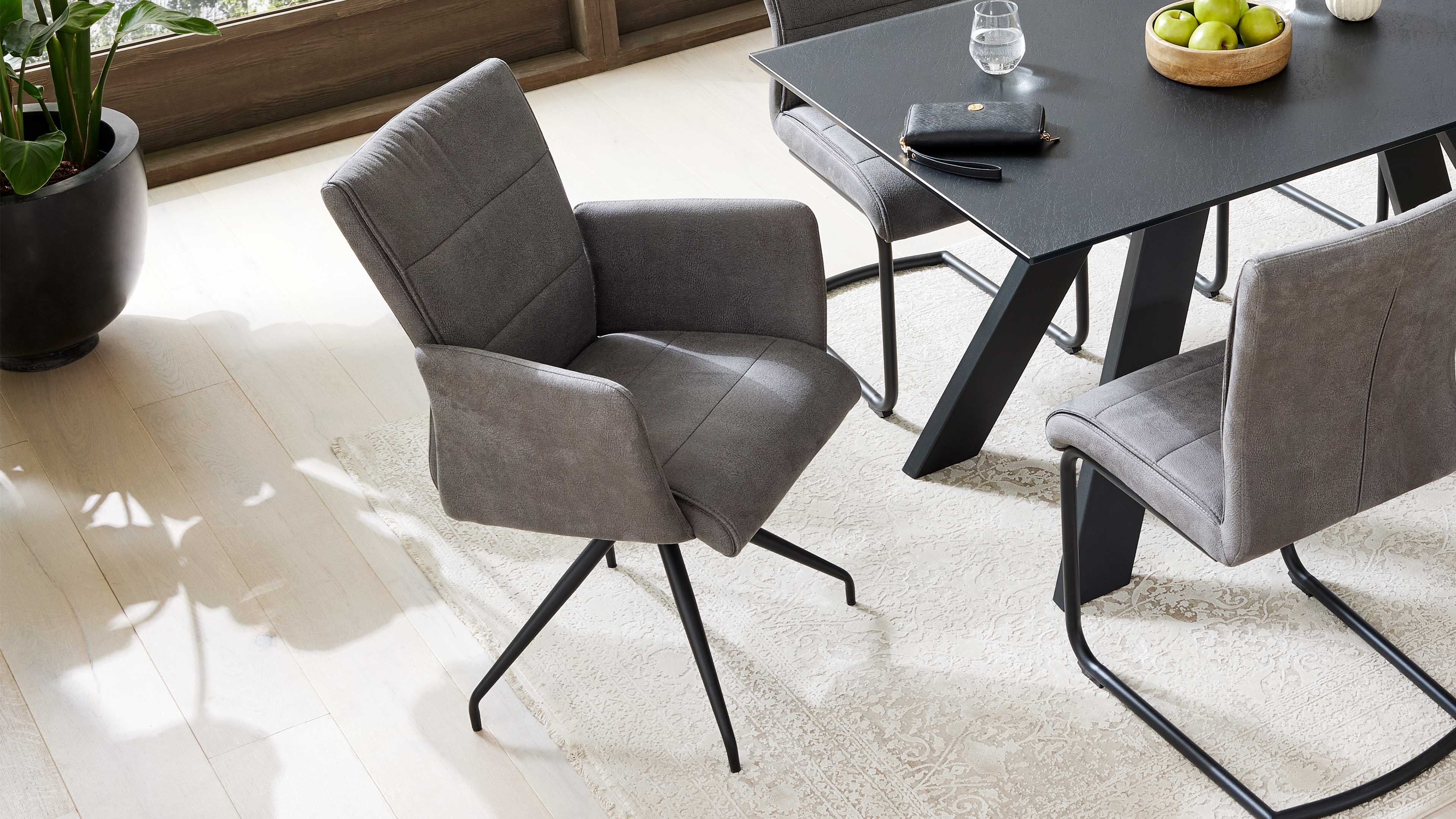 Grauer Polsterarmstuhl mit schwarzem vierarmigem Metallfuß vor schwarzem Esstisch mit Holzschale grüner Äpfel und weiteren grauen Stühlen.