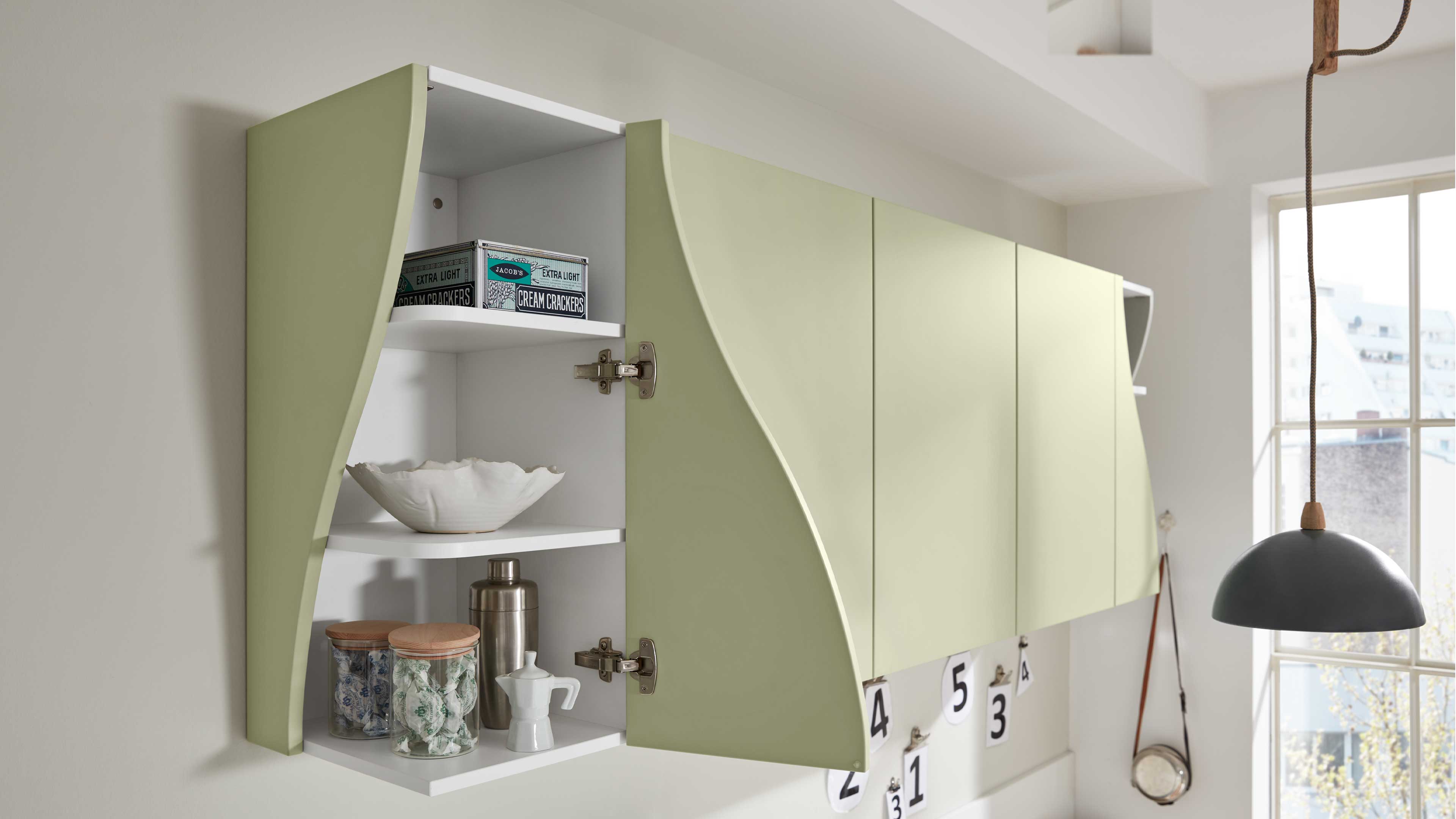 An der Wand hängender Küchenoberschrank mit hellgrünen, geschwungenen Türen, eine Tür geöffnet, weiße Innenböden und Küchenutensilien.