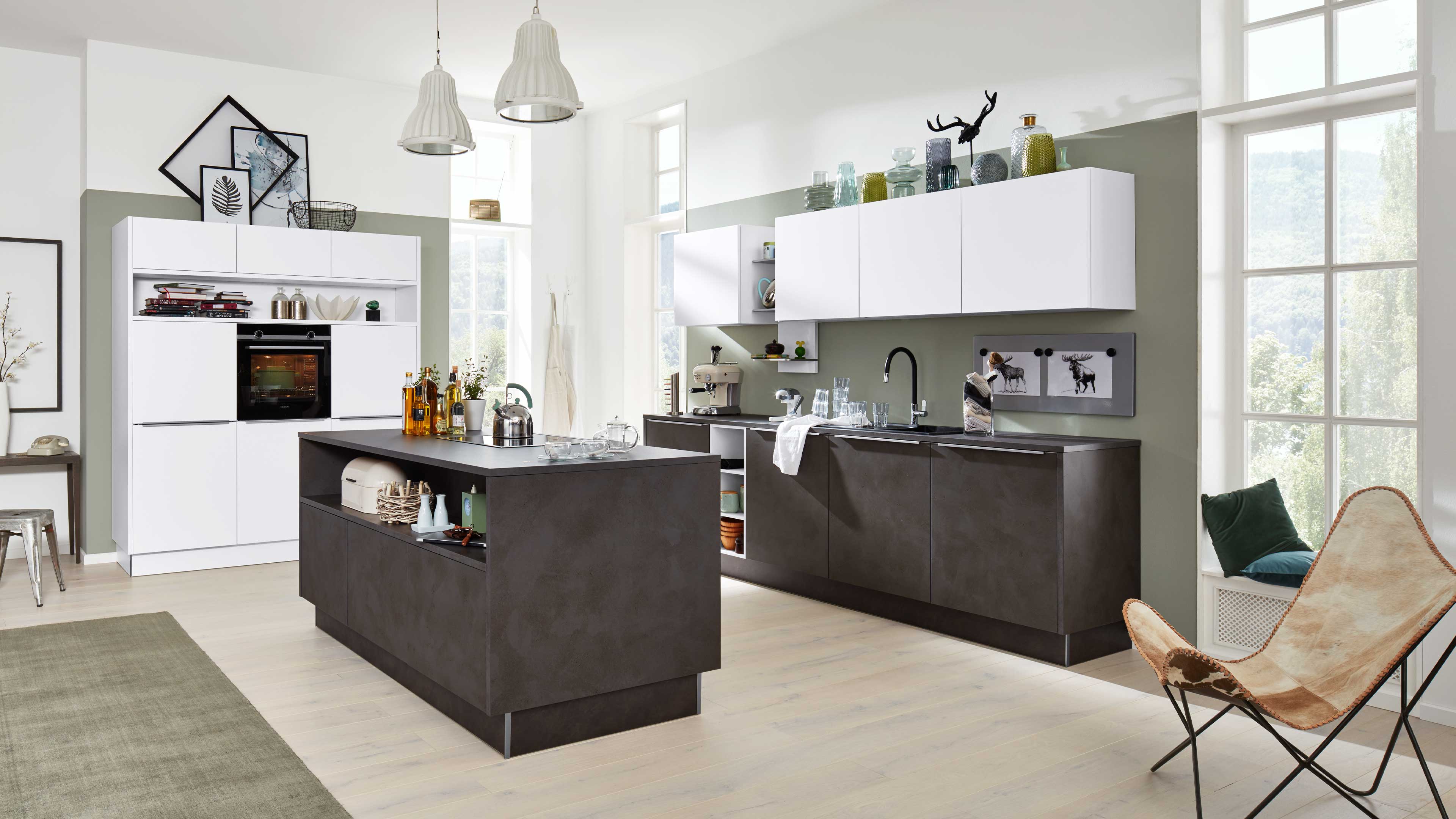 Offene Küche mit weißen Hängeschränken und dunkelgrauer Kücheninsel sowie Unterschränken in Betonoptik, großen Fenstern.