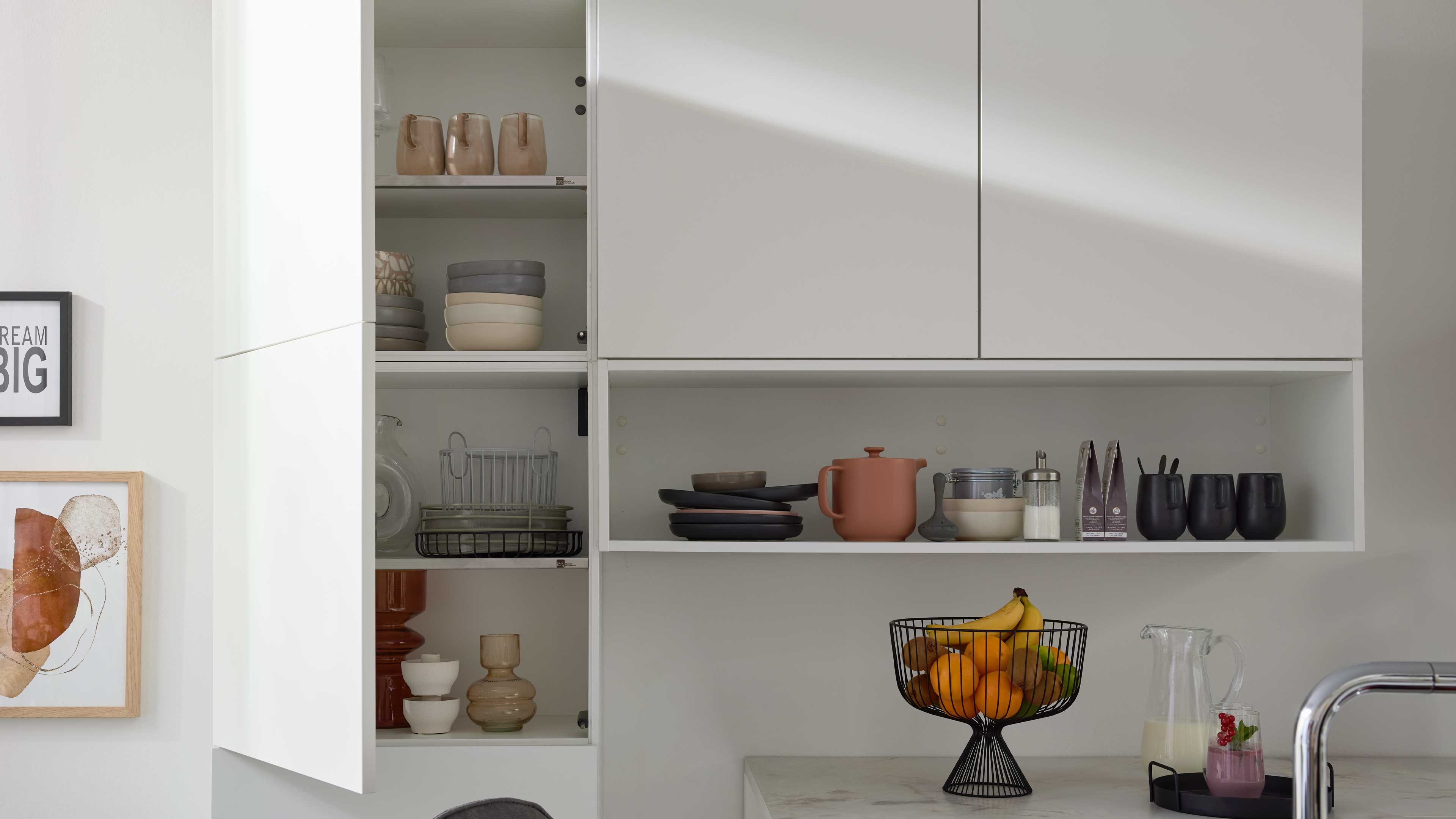 Weiße Küchenhängeschränke mit offenem Fach, darin Teller, Teekanne und schwarze Becher; darunter Obstkorb mit Bananen und Orangen.