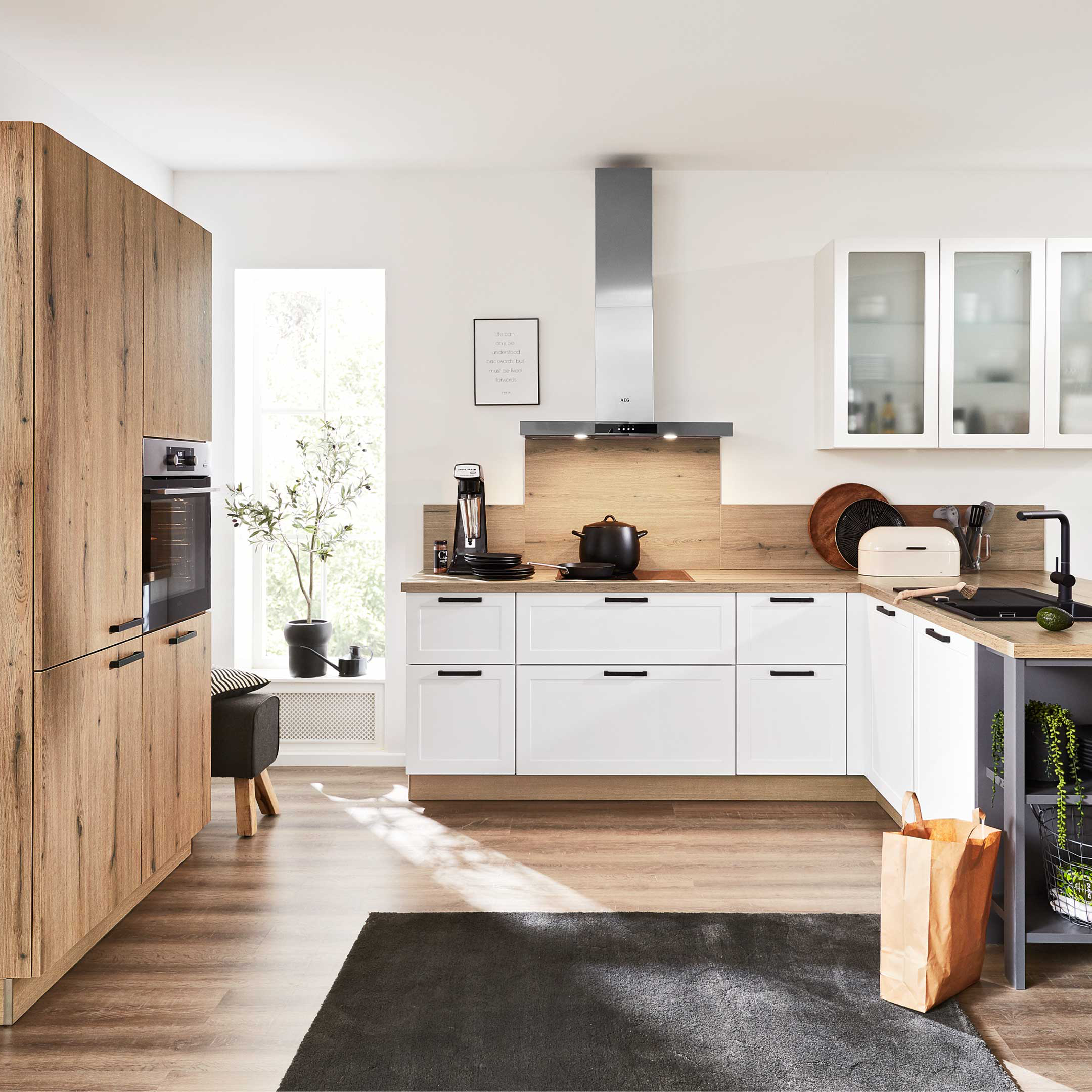 Küchenzeile mit Holz-Hochschrank links, weißen Unterschränken und Eckinsel mit offenen Regalen, Arbeitsplatte und Deko.