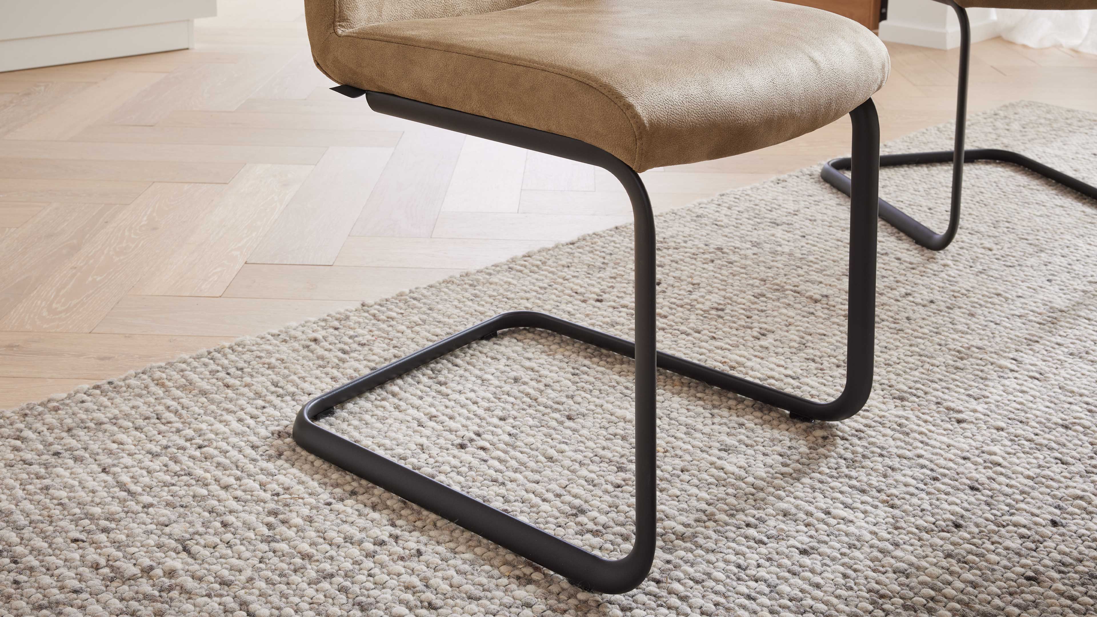 Hellbrauner gepolsterter Stuhl mit schwarzem freischwingendem Metallgestell auf grobem hellbeigem Teppich vor hellem Fischgrätparkett.