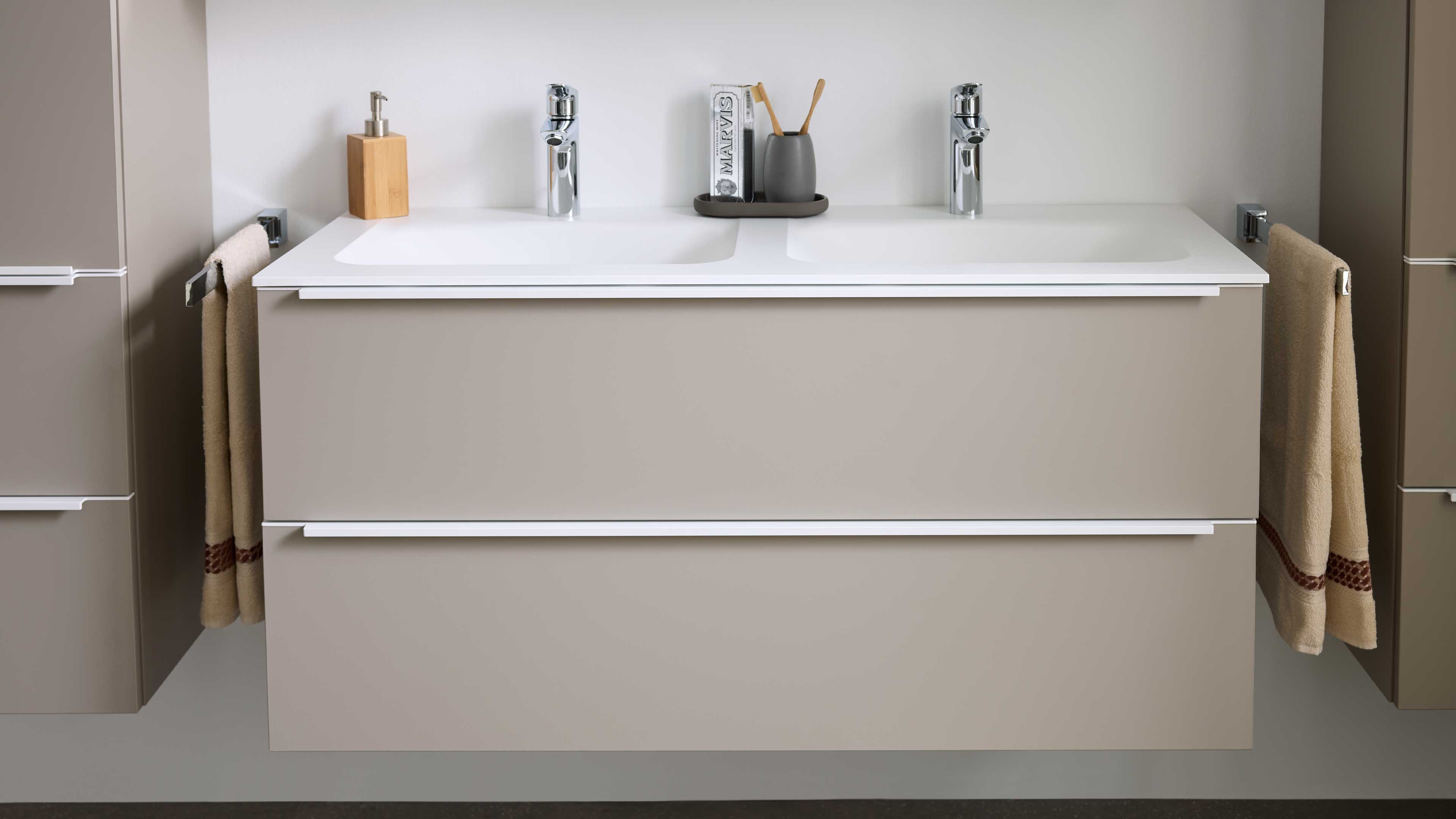 Hängender Doppelwaschtisch mit glatter beiger Front, weißer Aufsatzplatte, zwei verchromten Armaturen und beigen Handtüchern an den Seiten.