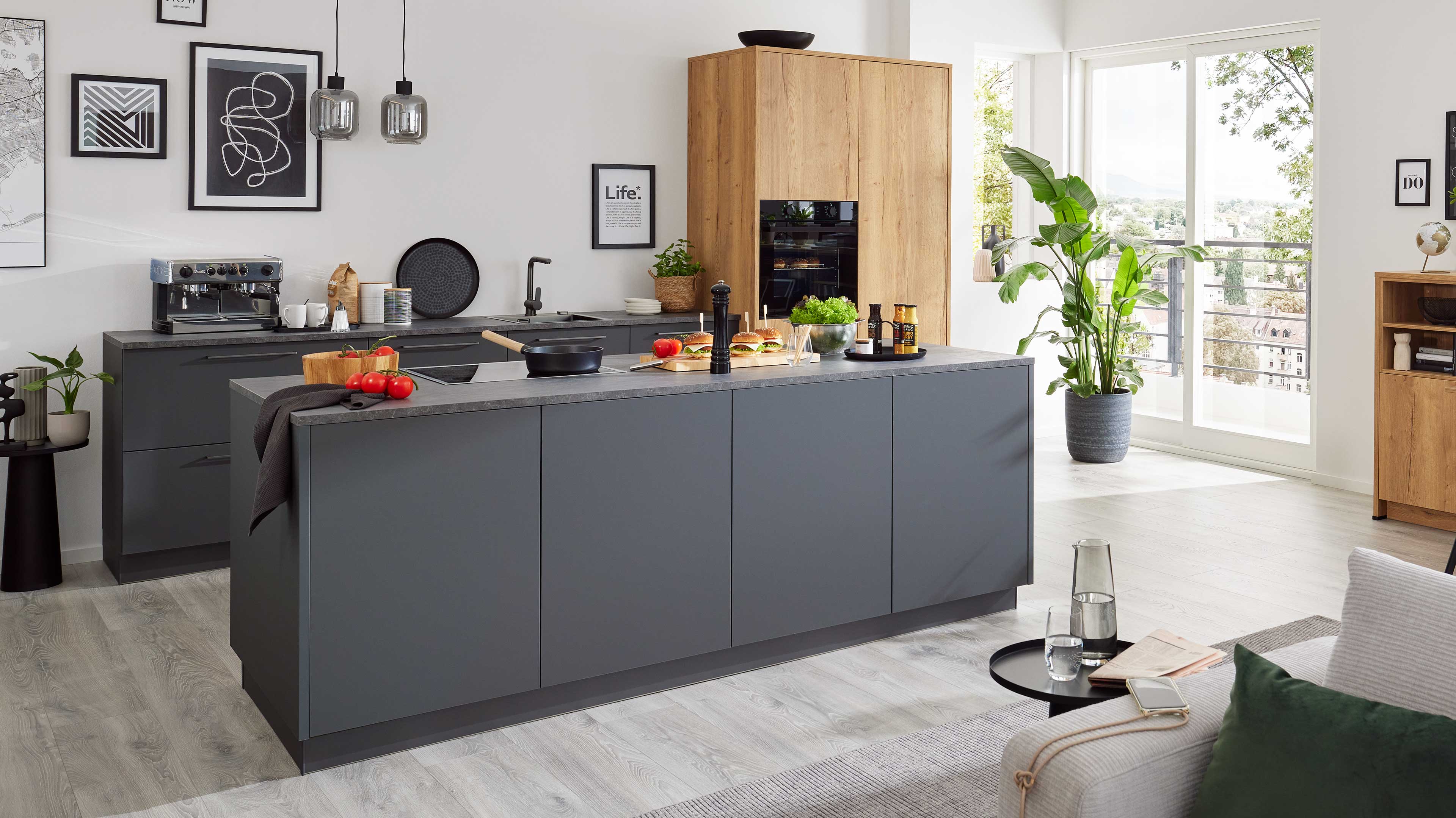 Große dunkle Kücheninsel mit grauer Arbeitsplatte, Kochfeld, Schneidebrett mit Tomaten und daneben Holzhochschrank und Balkonfenster.