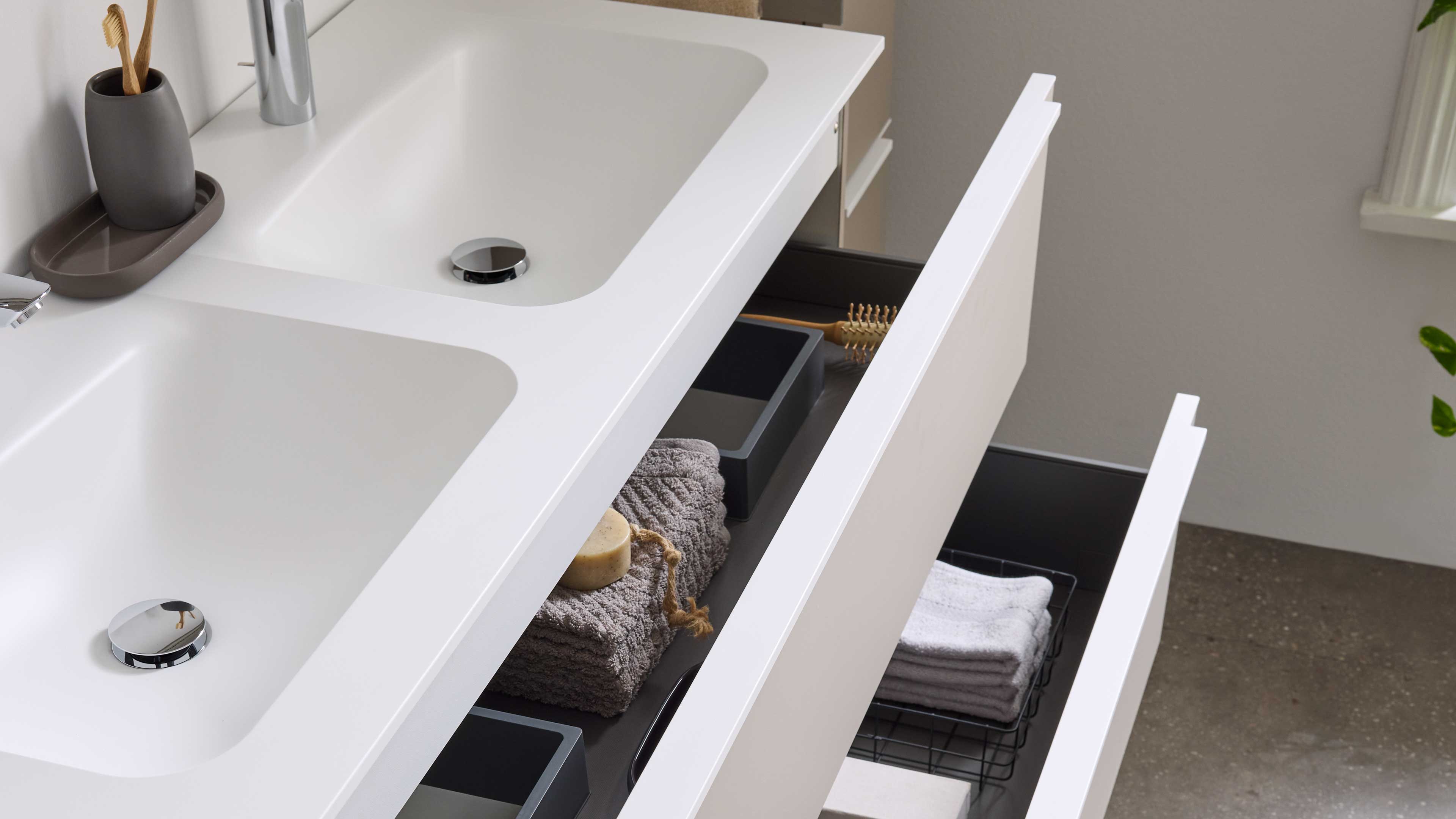 Weißer Waschtisch mit Doppelbecken und zwei offenen Schubladen, darin graue Handtücher, Seife, Bürsten und kleine Boxen.
