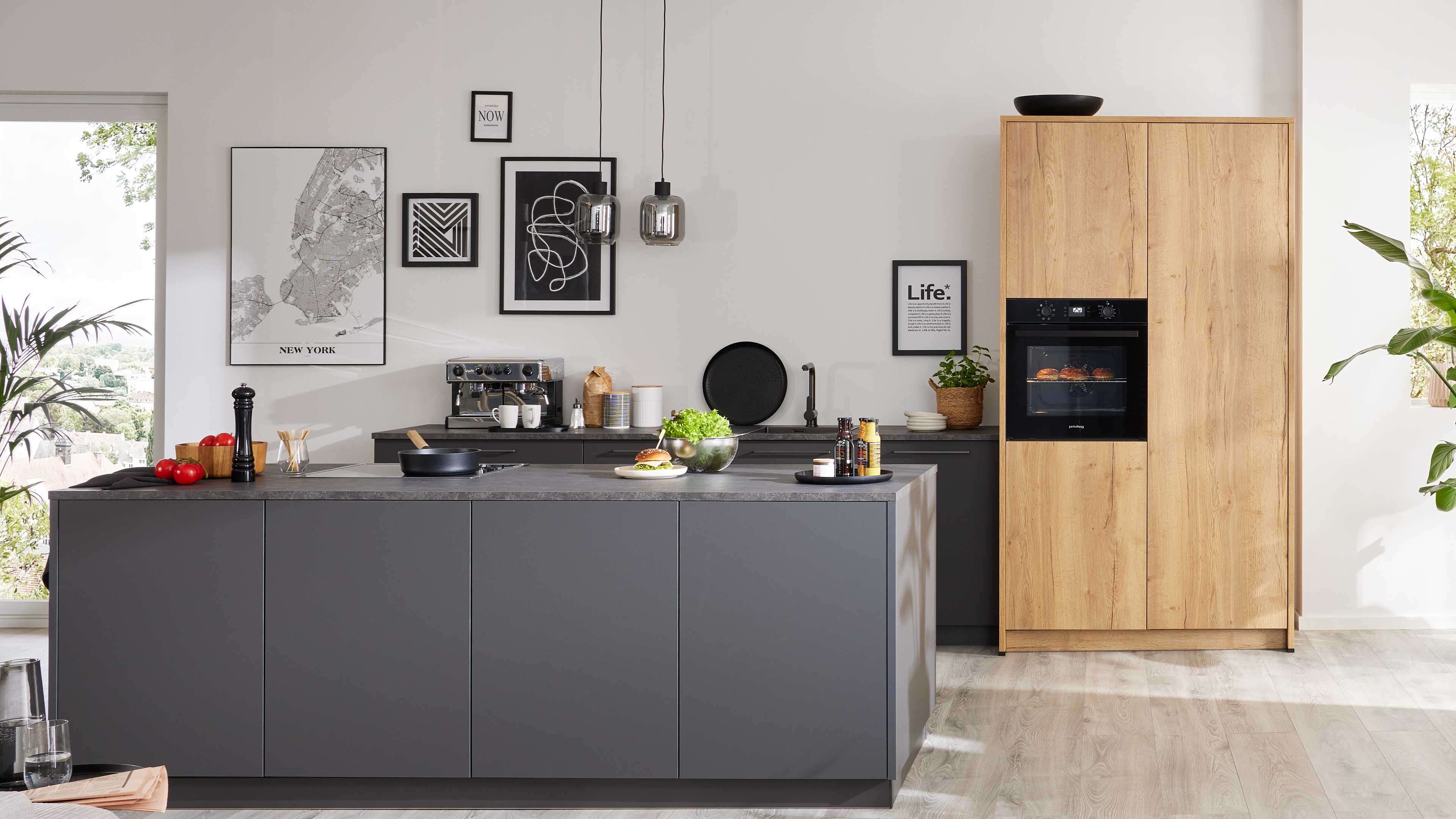 Dunkelgraue Kücheninsel mit Arbeitsfläche, Pfanne, Pfeffermühle und Salatschüssel; dahinter schwarze Schränke und Holzschrankwand mit Ofen.