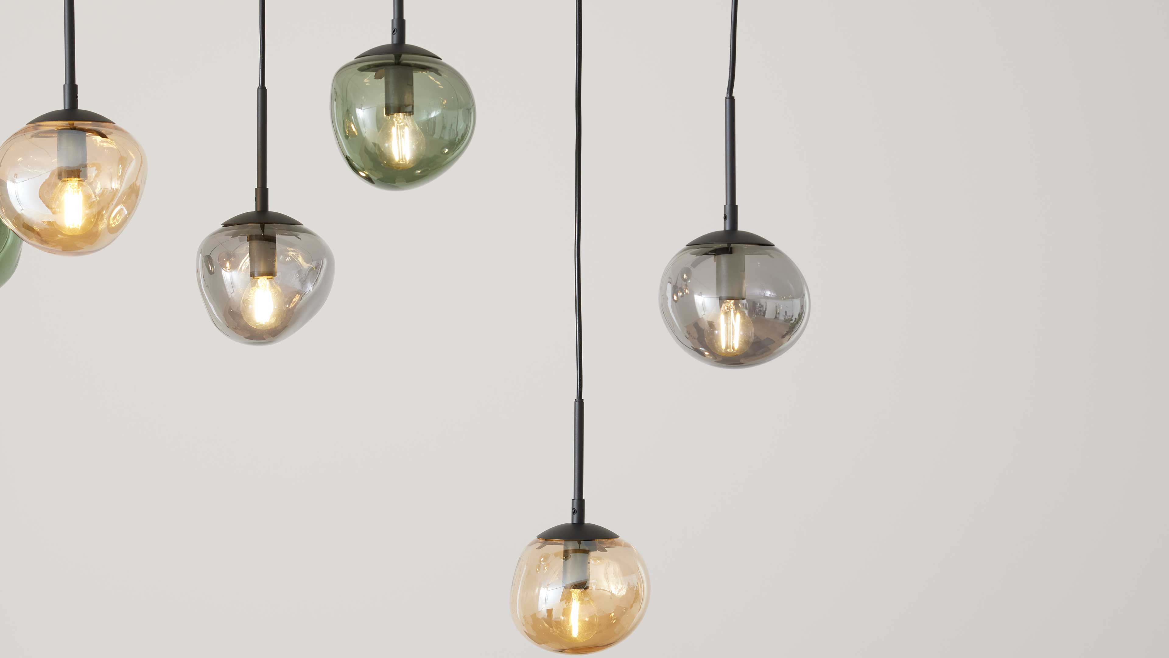 Fünf hängende Glas-Pendelleuchten in bernstein-, grün- und rauchfarbenen Kugeln mit schwarzen Kabeln vor heller Wand.