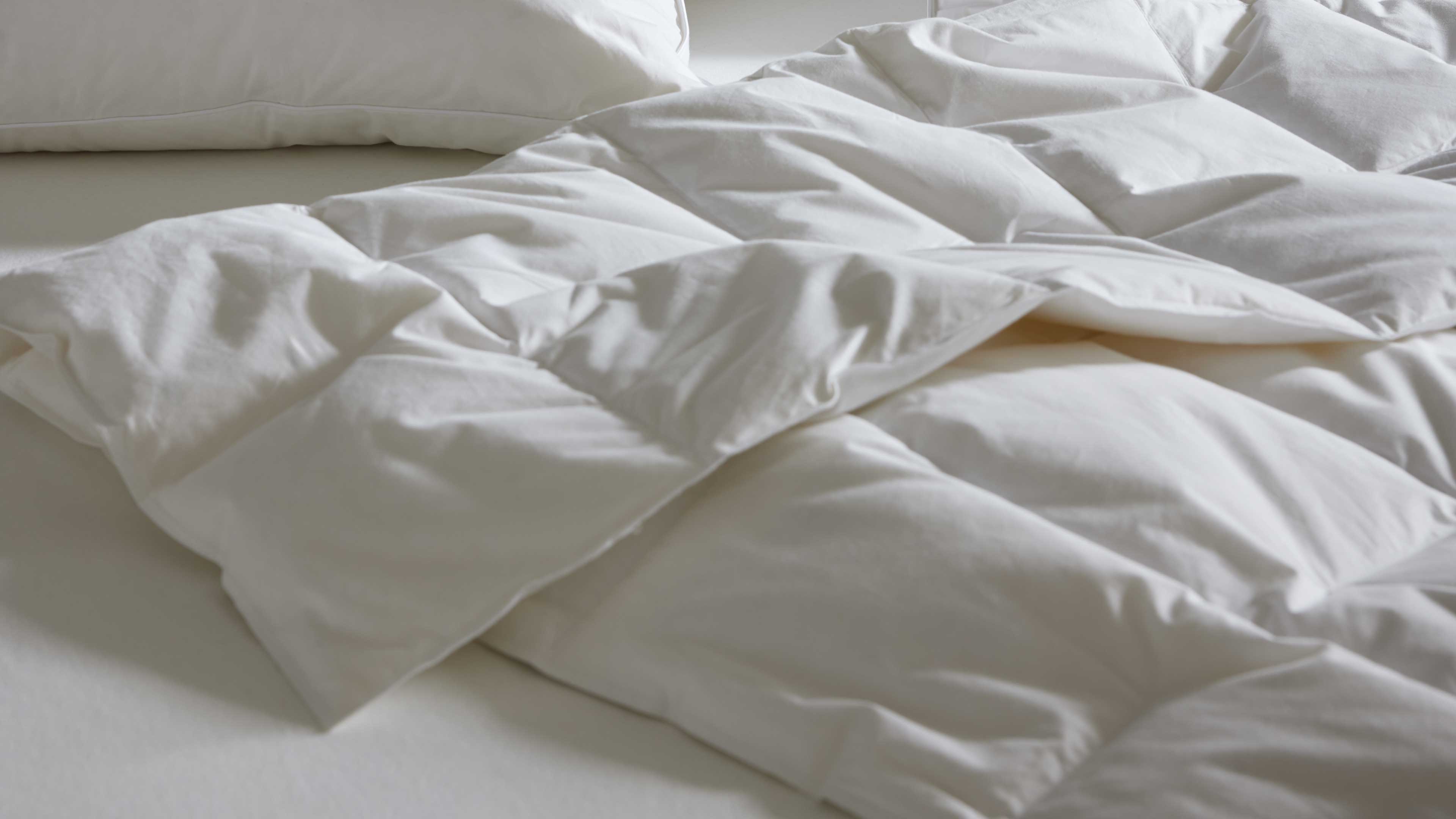 Weiße gestepptete Decke liegt zerwühlt auf Matratze, im Hintergrund ist der Rand eines weißen Kissens zu sehen.