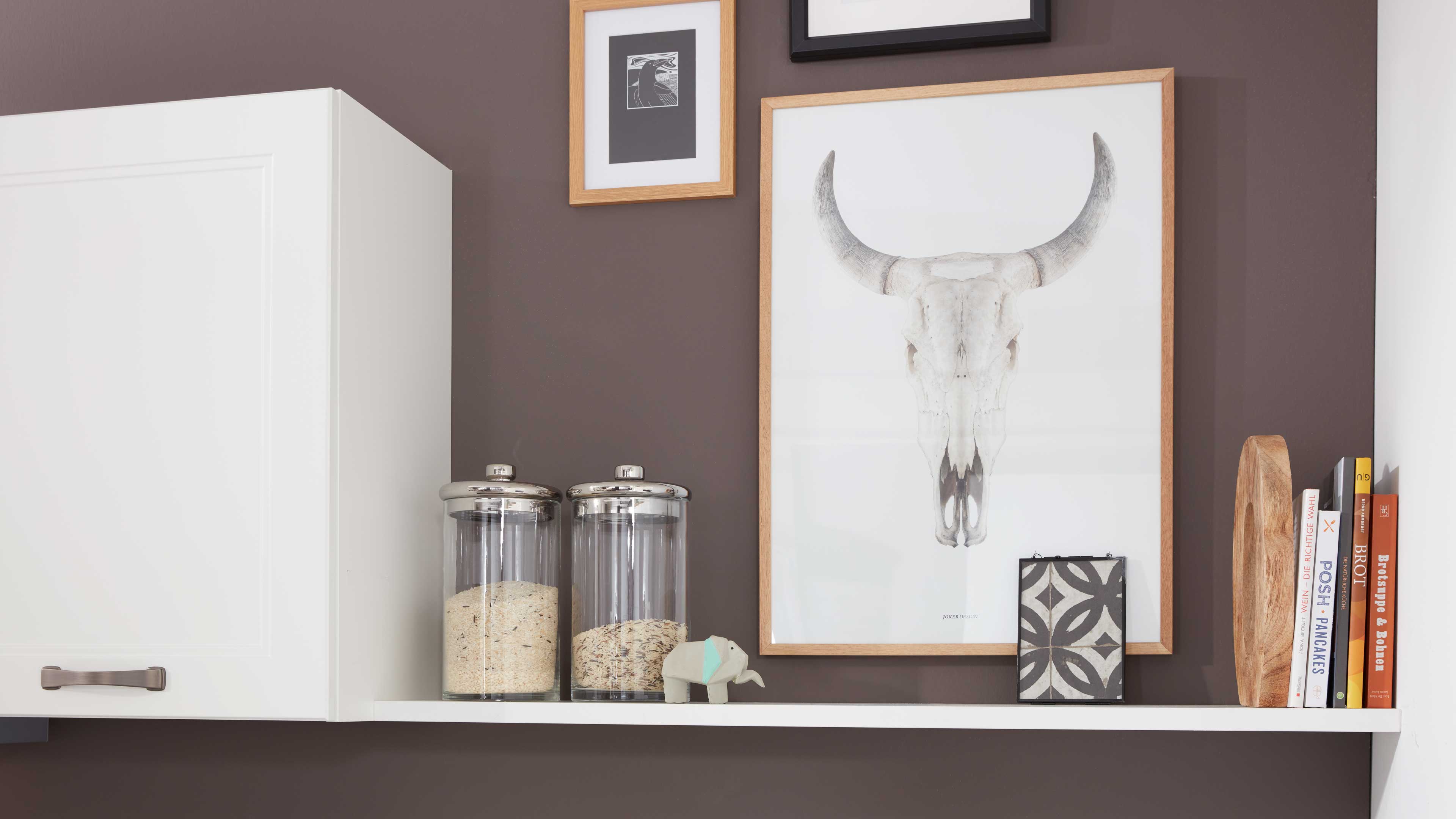 Weißes Wandregal mit zwei Glasvorratsgläsern mit Metalldeckeln, kleinem Elefanten und großem Bild mit Schädel in Holzrahmen vor brauner Wand.