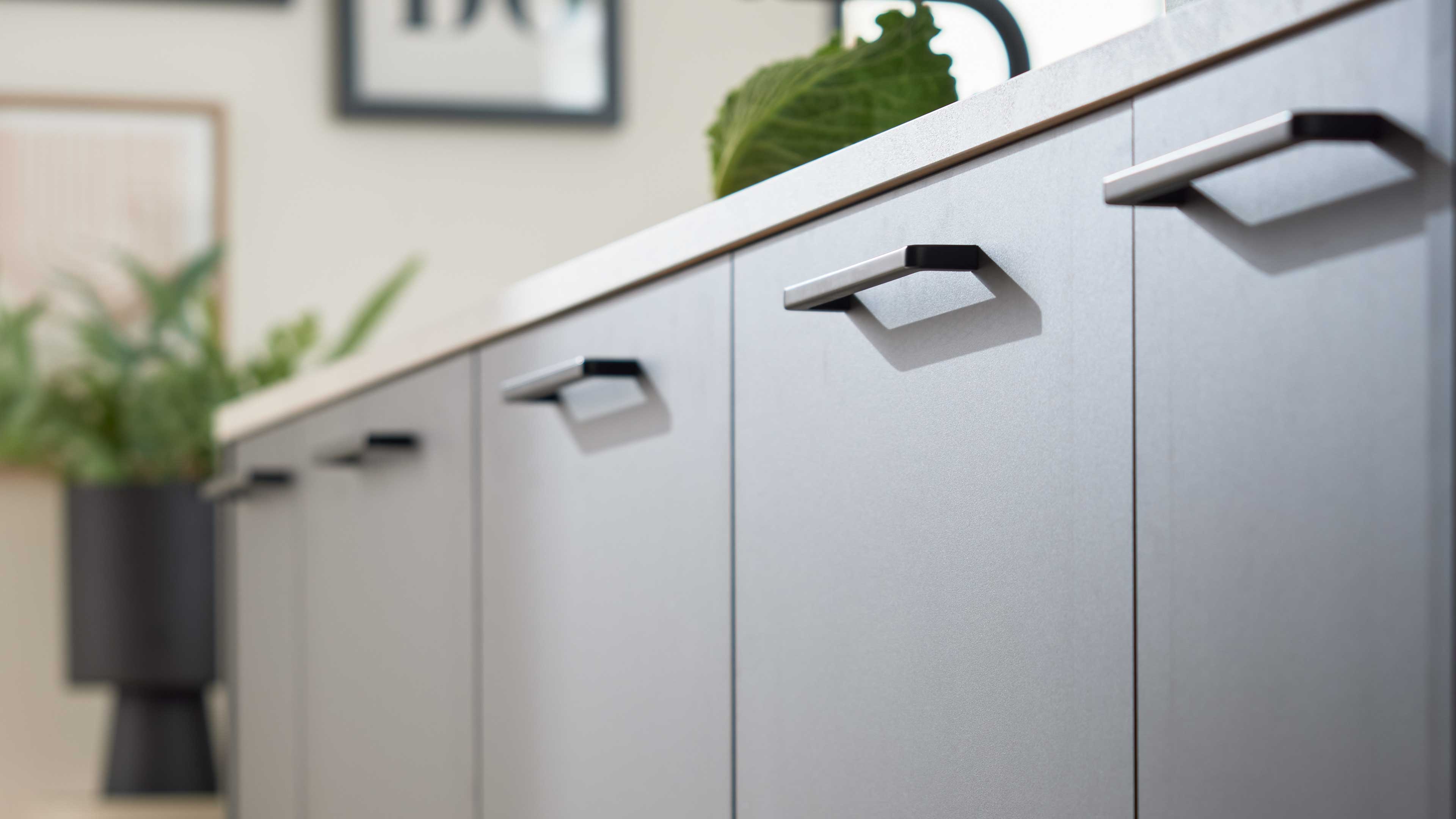 Graue Schrankfronten mit schmalen silbernen Griffen in Reihe, oben helle Arbeitsplatte und verschwommene Zimmerpflanze im Hintergrund.