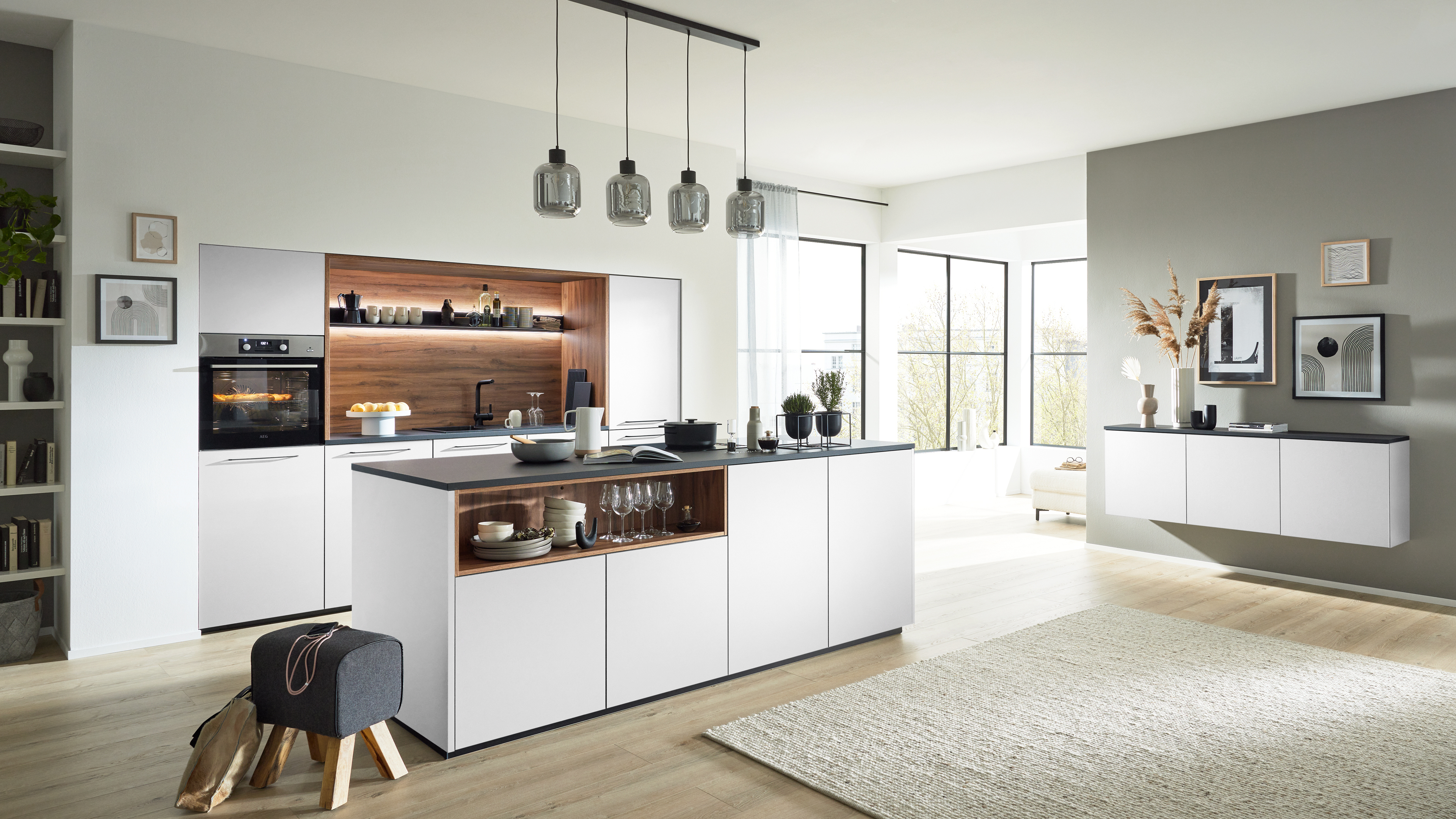 Weiße Kücheninsel mit dunkler Arbeitsplatte und offenem Holzfach mit Geschirr; im Hintergrund Einbauherd, Nische und hängende Glaslampen.