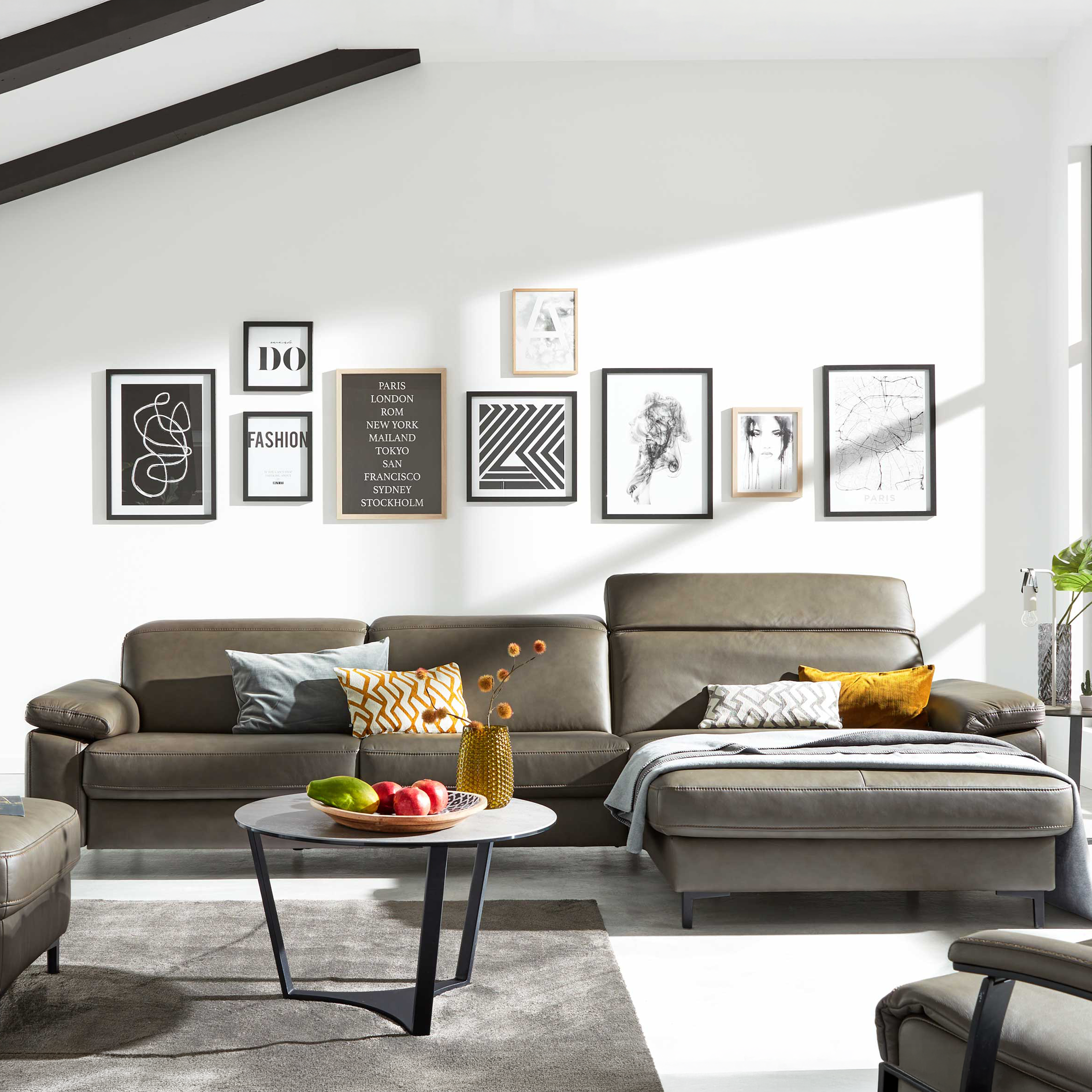 Grau-braunes Ecksofa mit Chaiselongue, Kissen und Decke vor Wand mit neun gerahmten Bildern und großem Fenster.