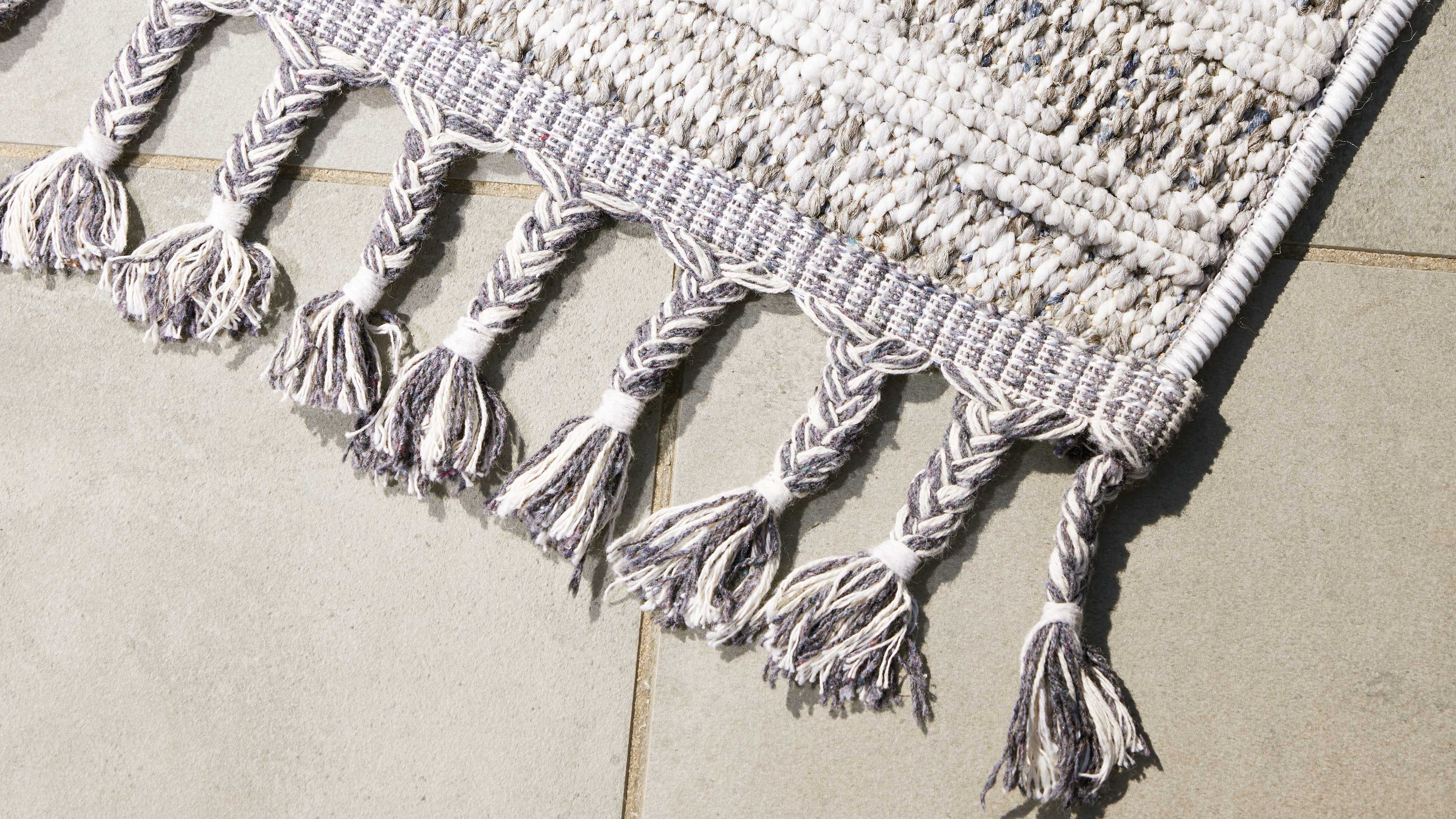 Gewebter Teppichrand mit abwechselnden geflochtenen Quasten in Creme und Grau liegt auf hellgrauen Bodenfliesen.