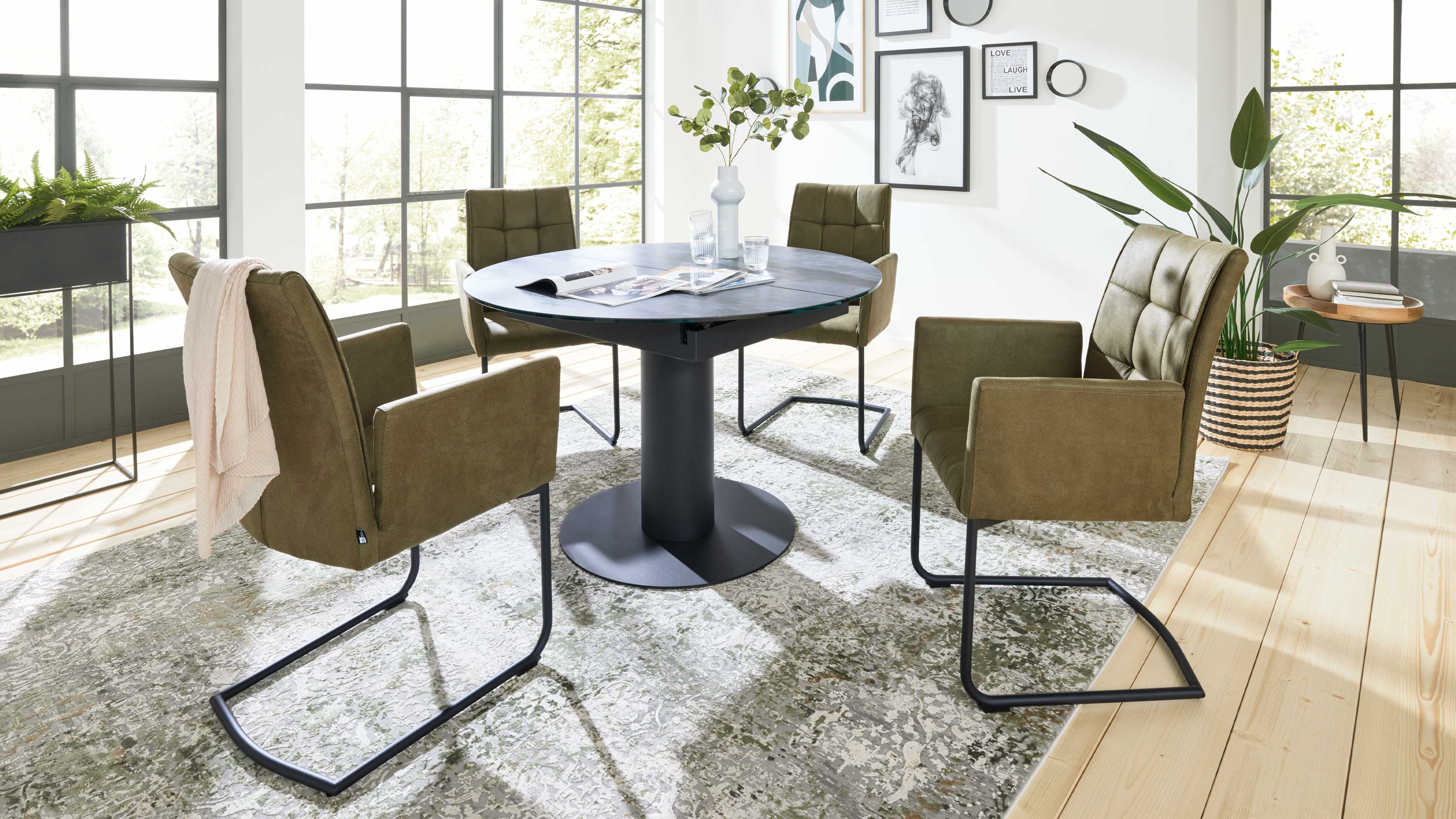 Runder schwarzer Esstisch mit Vase, umgeben von vier olivgrünen Stühlen in hellem Raum vor großen Fenstern, Teppich und Holzboden.