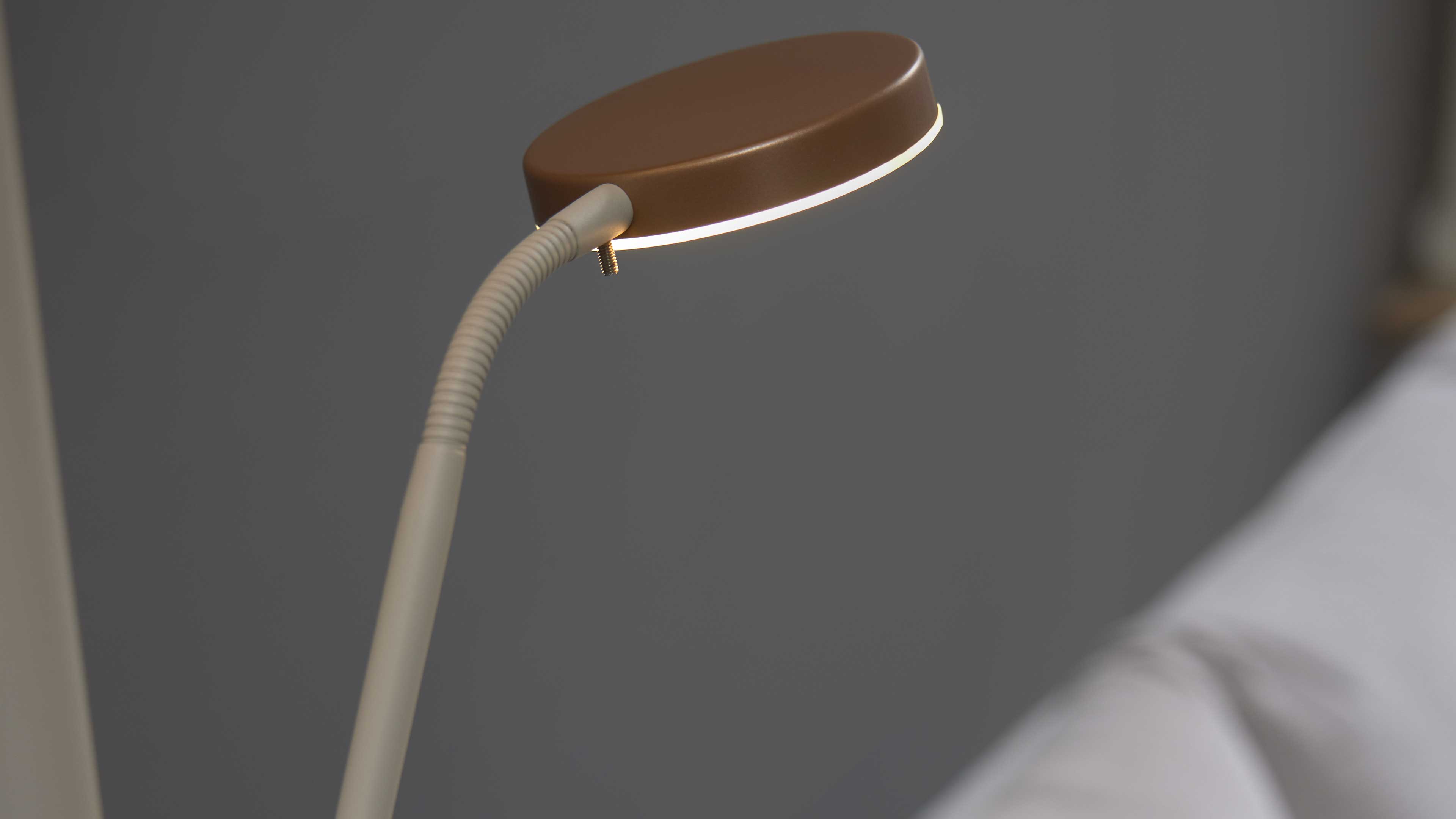 Flexible Leselampe mit rundem braunem Lampenschirm und weißem geriffeltem Schwanenhals, eingeschaltetem Licht vor grauem Hintergrund.