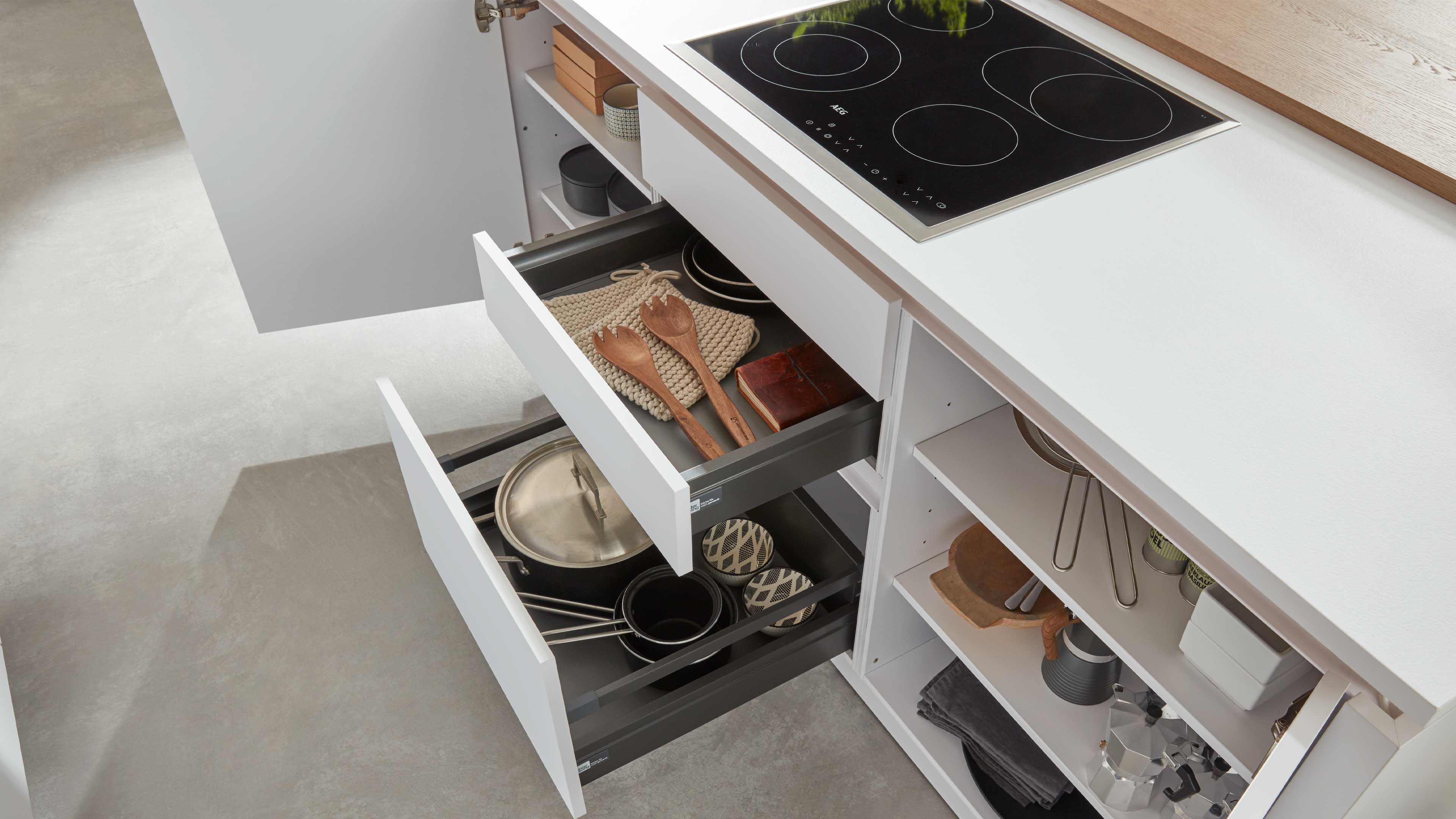 Weißer Küchenunterschrank mit zwei geöffneten Schubladen, dunklen Einsätzen und oben schwarzem Kochfeld, Holzlöffel und Töpfe sichtbar.