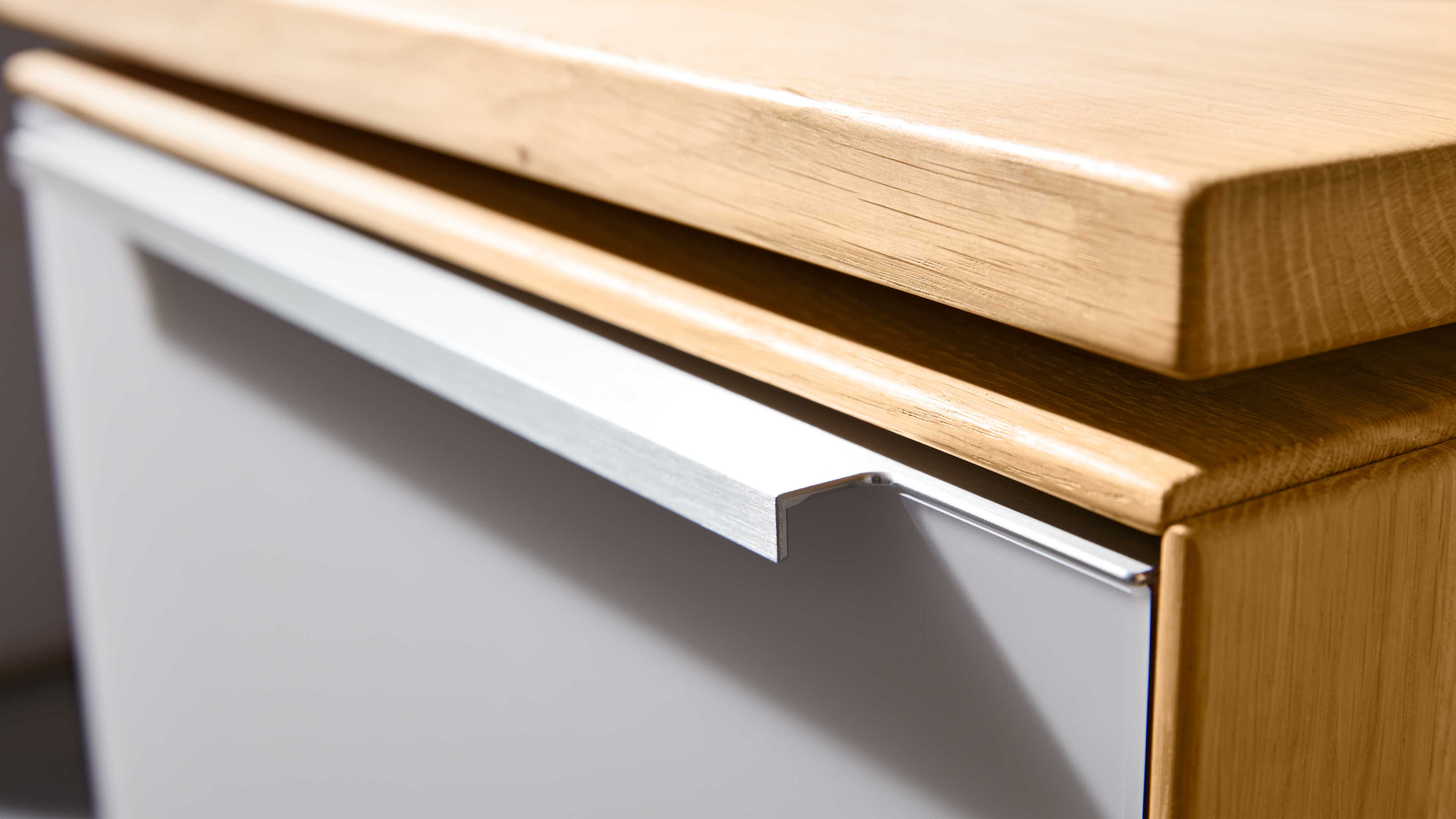 Detailaufnahme einer hellen Holzarbeitsplatte mit abgerundeter Kante, darunter weiße Schubladenfront und silberner Griffleiste.