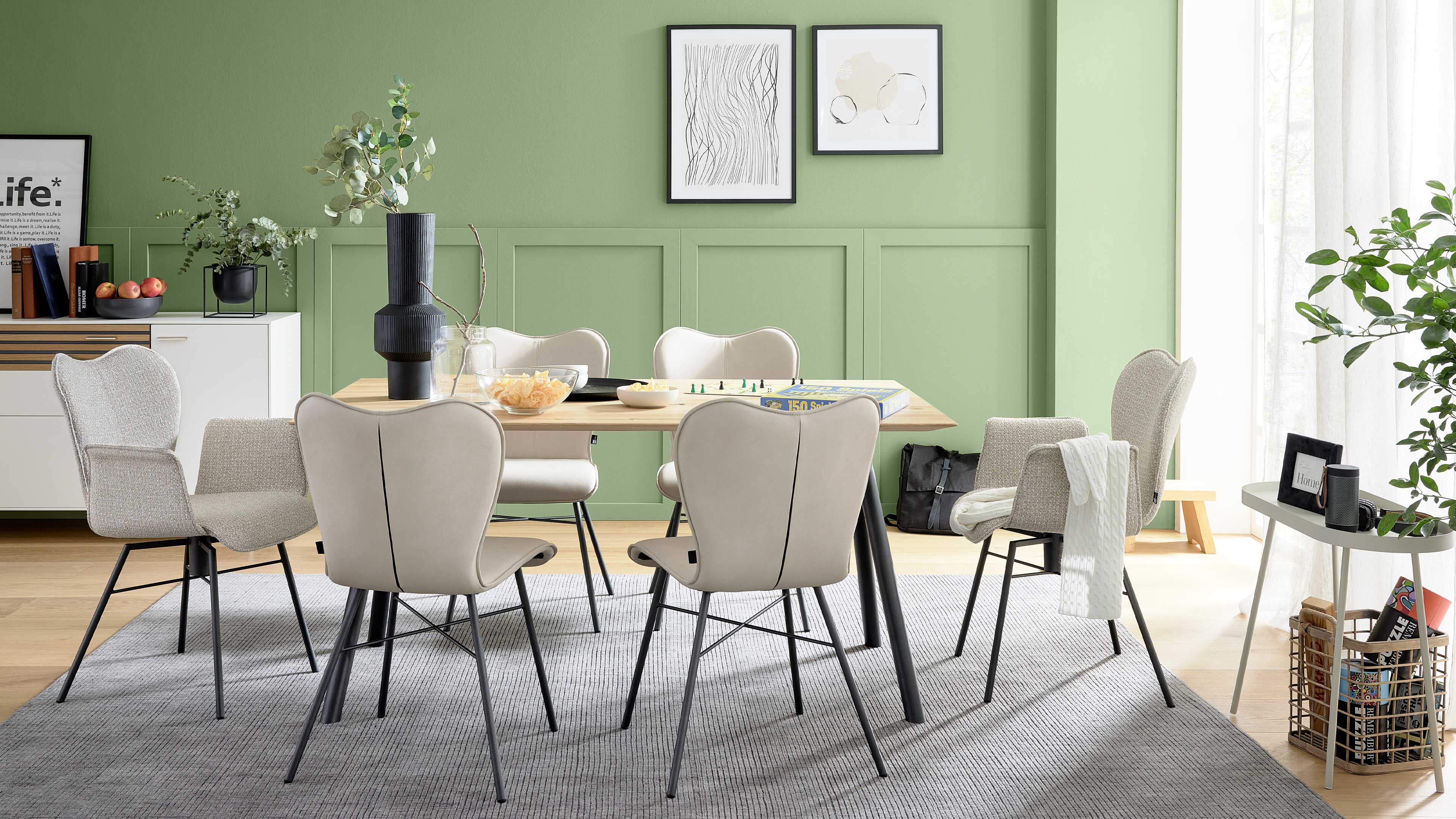 Esstisch aus hellem Holz mit sechs hellbeigen Polsterstühlen auf grauem Teppich vor grün getäfelter Wand mit zwei Bilderrahmen.