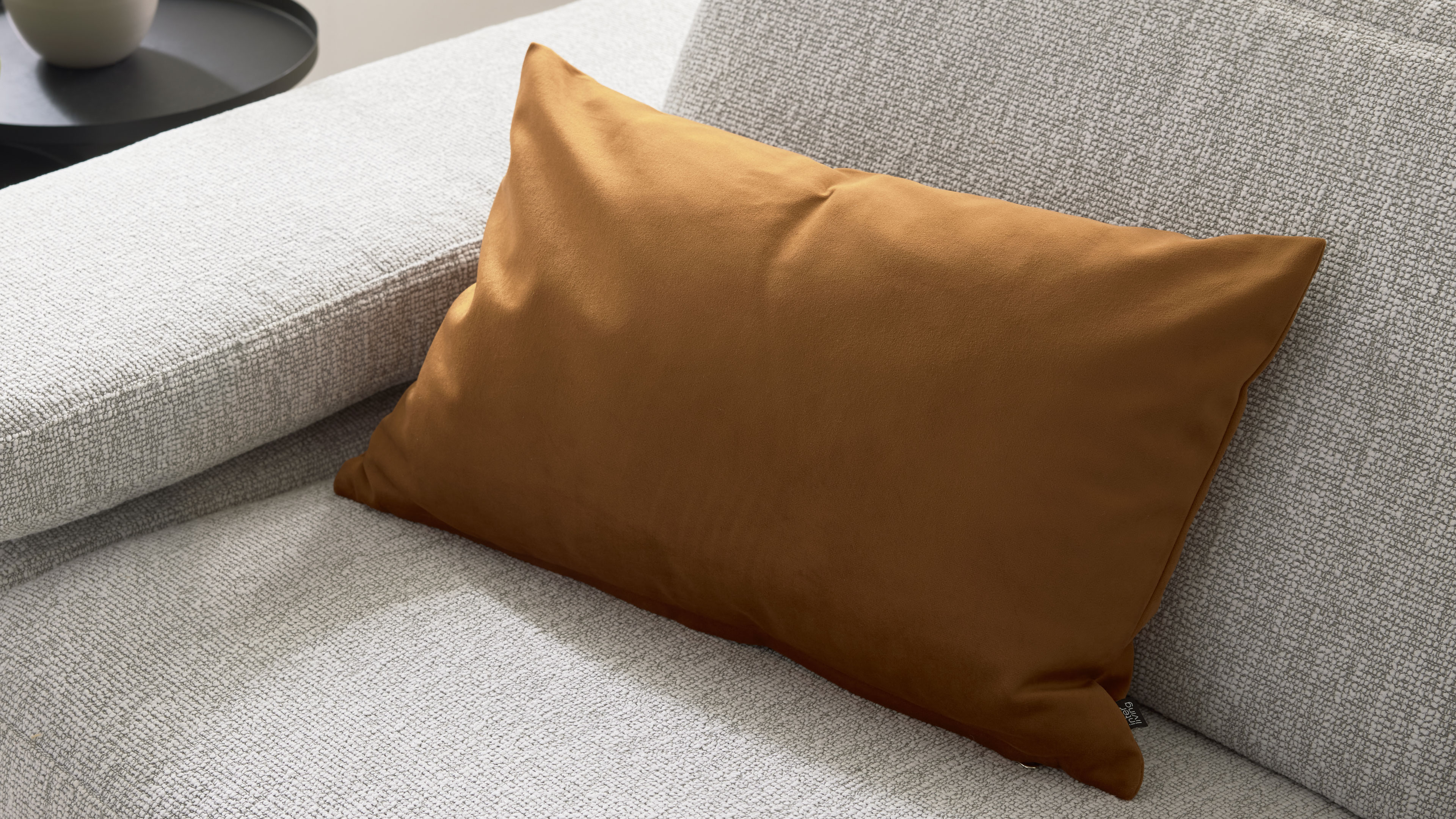Rechteckiges, rostbraunes Zierkissen liegt auf hellgrauem, strukturiertem Sofa, links oben ein schwarzer Beistelltisch mit Vase.