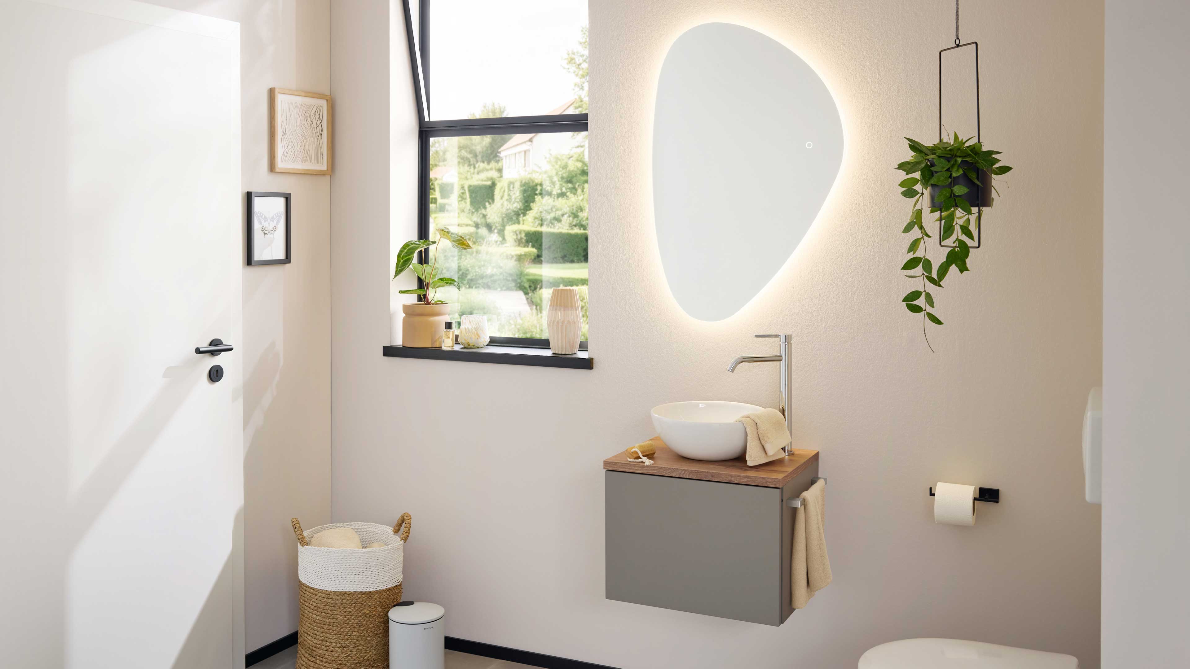 Schwebender Waschtisch mit grauer Front, Holzplatte, weißer Aufsatzwaschschale, Armatur und organisch geformtem, hinterleuchtetem Spiegel.