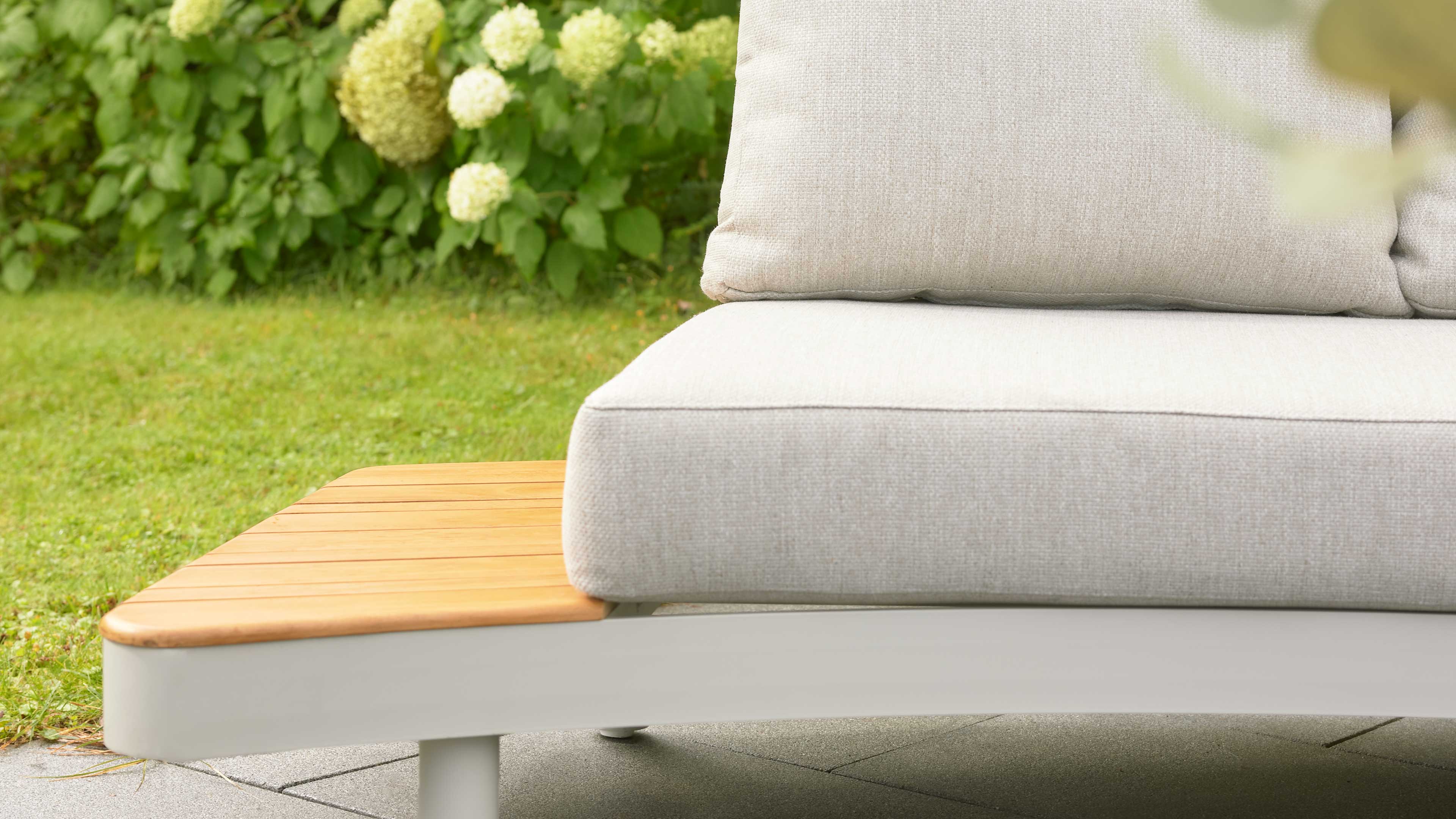 Rechte Sitzfläche eines hellgrauen Outdoor-Sofas mit dickem Sitzkissen und Holzablage links, im Hintergrund grüner Rasen und weiße Blüten.