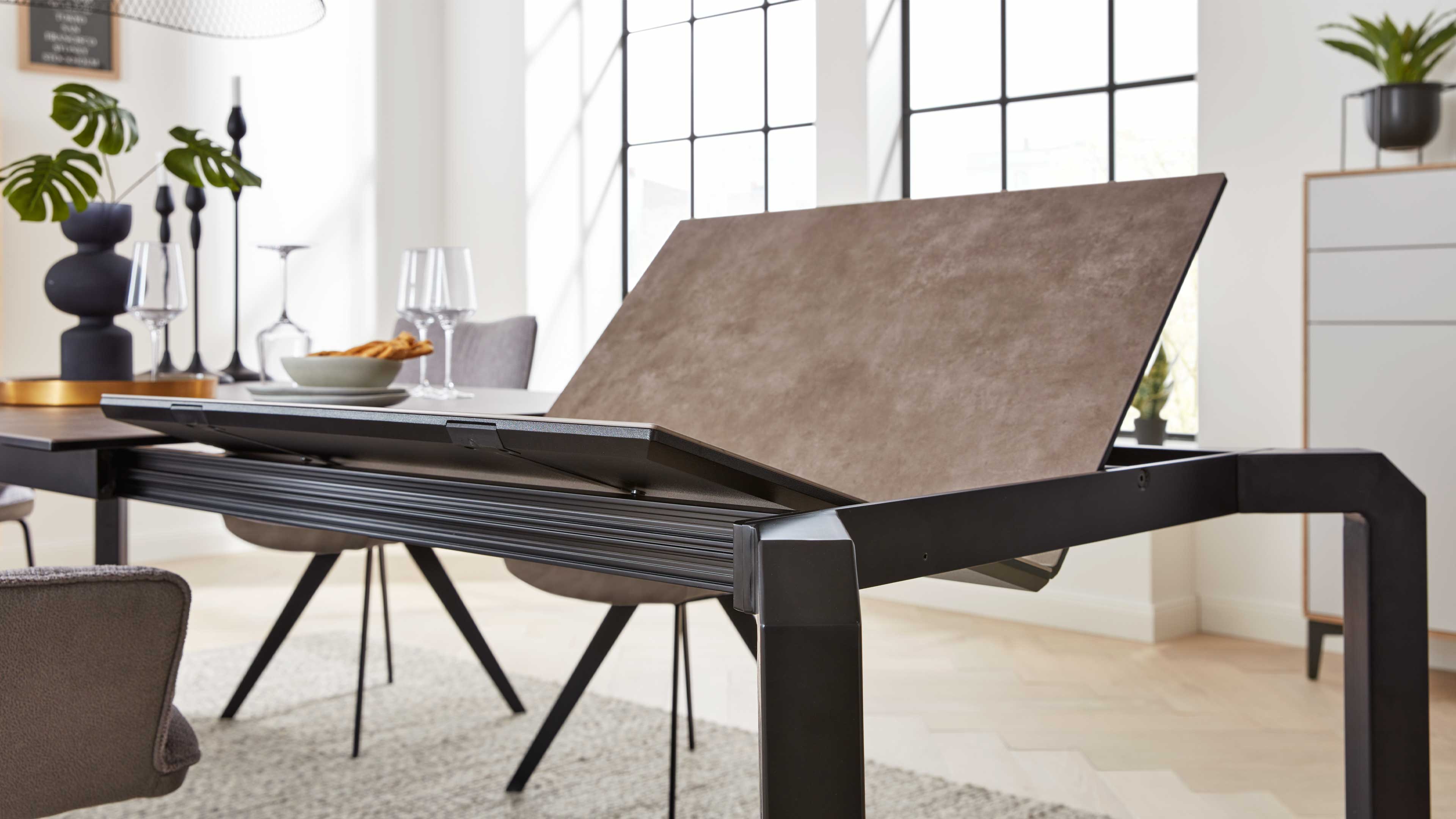 Moderner Esstisch mit schwarzem Metallgestell und hochgeklappter brauner Einlegeplatte, dahinter Stühle und Tischdeko.