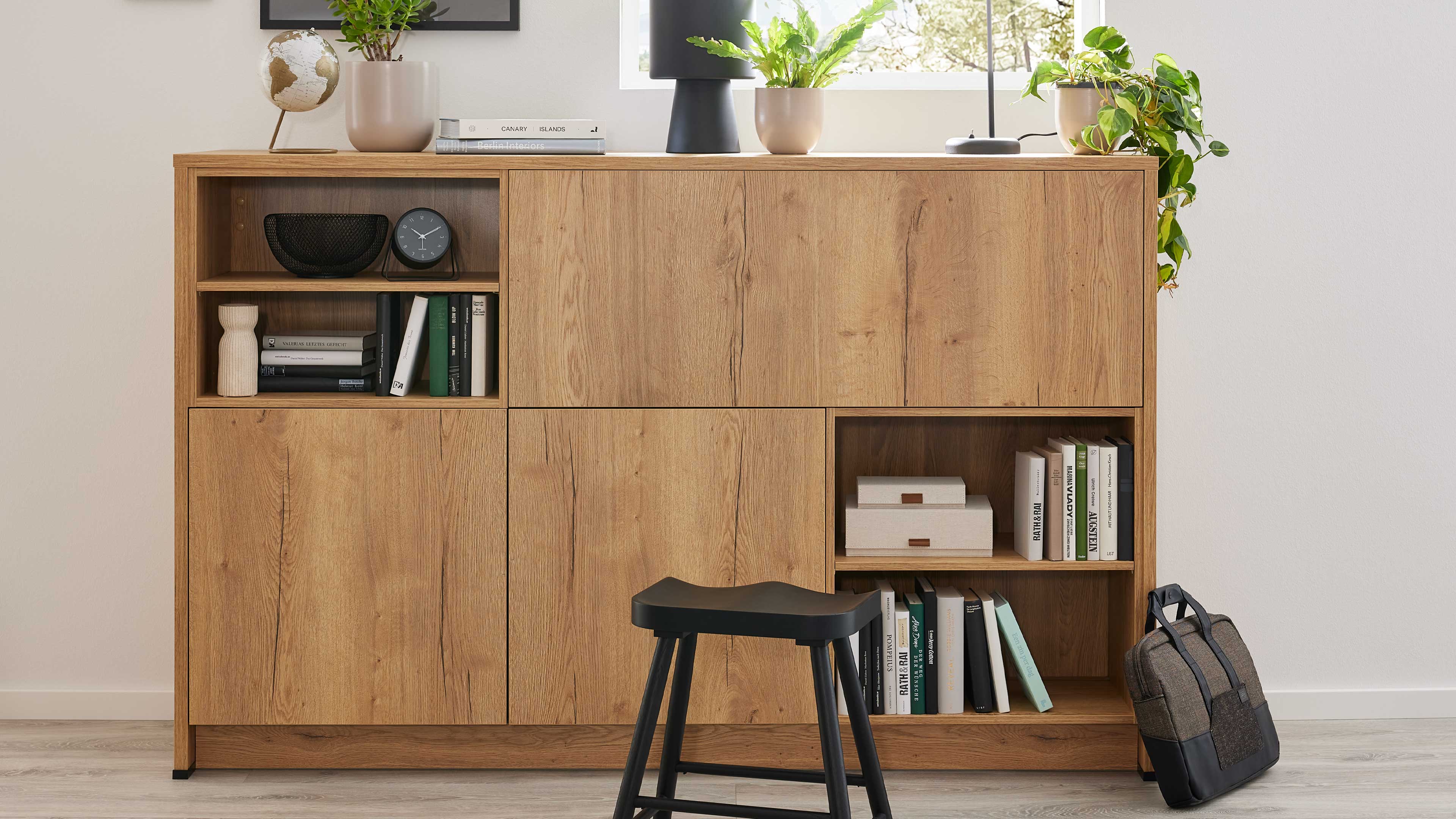 Helles Holzsideboard mit offenen Fächern und Türen, dekoriert mit Pflanzen, Lampe, Büchern; schwarzer Hocker vorne, Rucksack rechts.