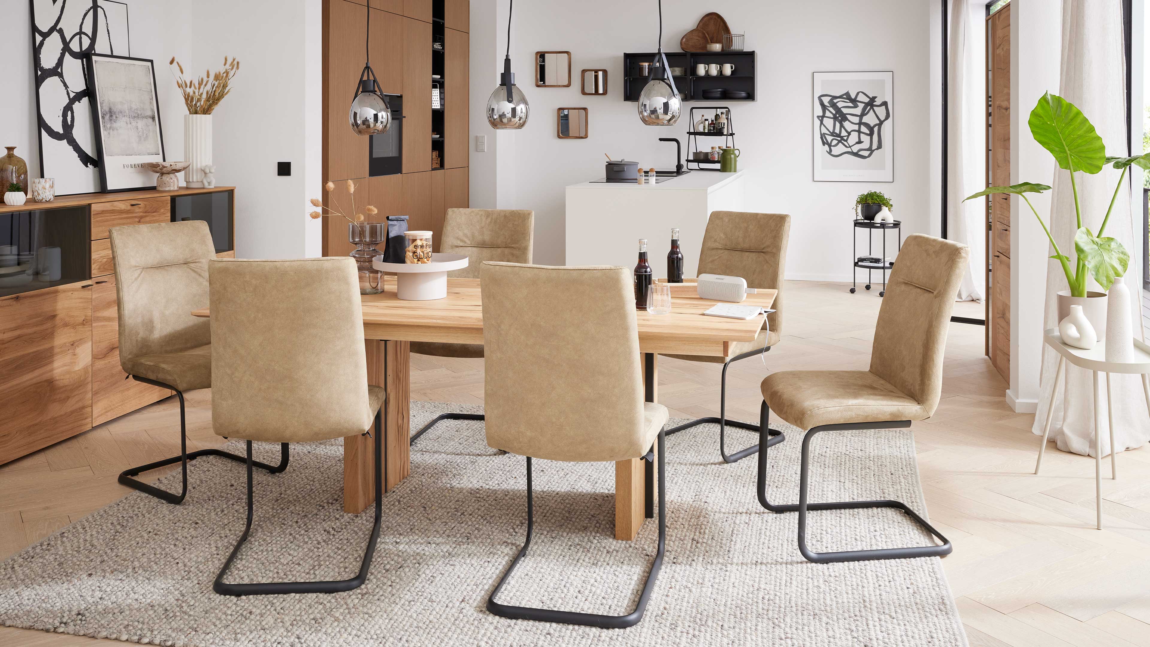 Rechteckiger Holztisch mit sechs beige gepolsterten Freischwinger-Stühlen, darauf Deko, vor Sideboard und Hängelampen.
