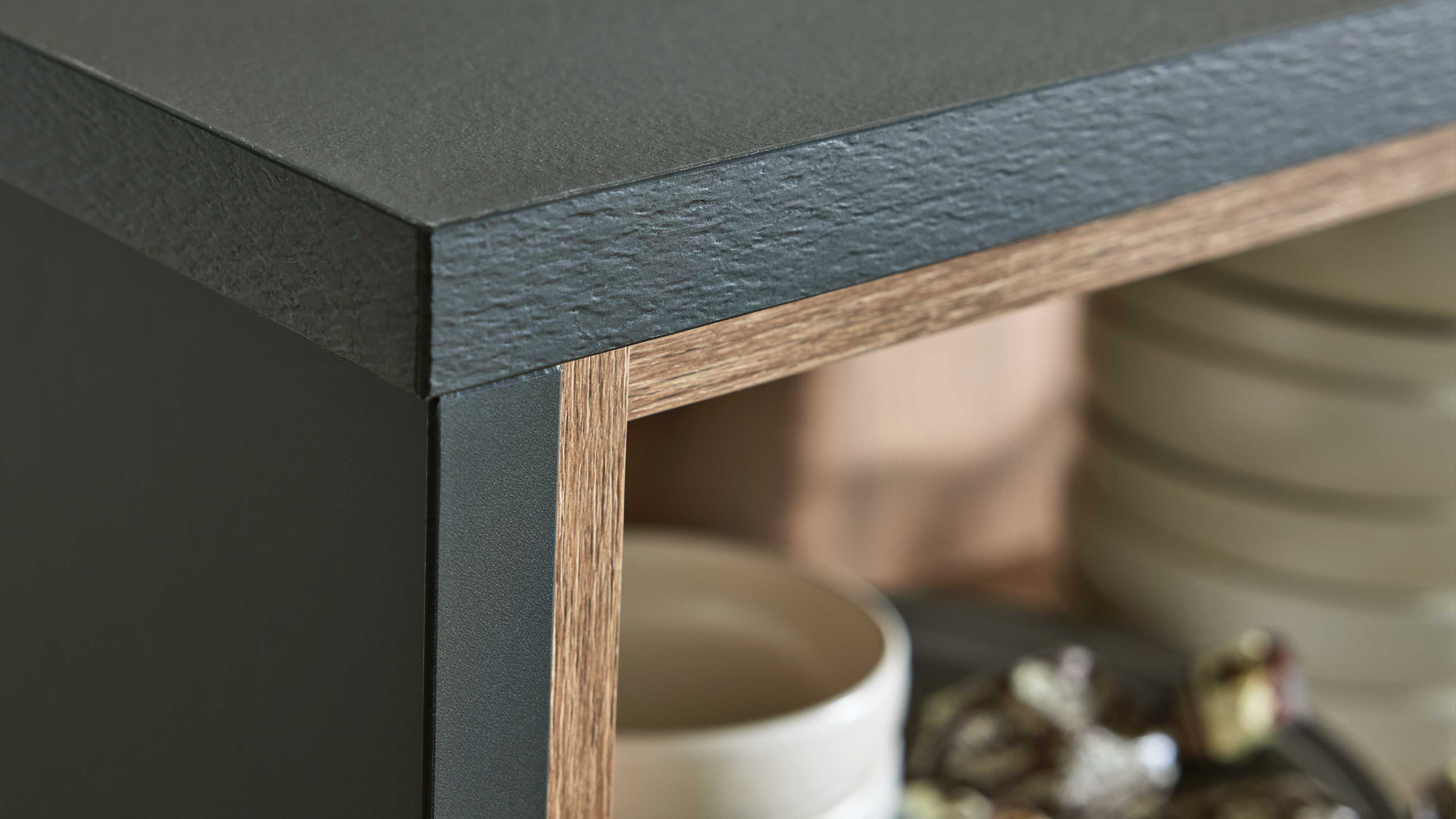 Eckansicht eines Schranks mit schwarzer, strukturierter Arbeitsplatte, schwarzer Seitenwand und Holzdekorrahmen, unscharfe Schalen im Fach.