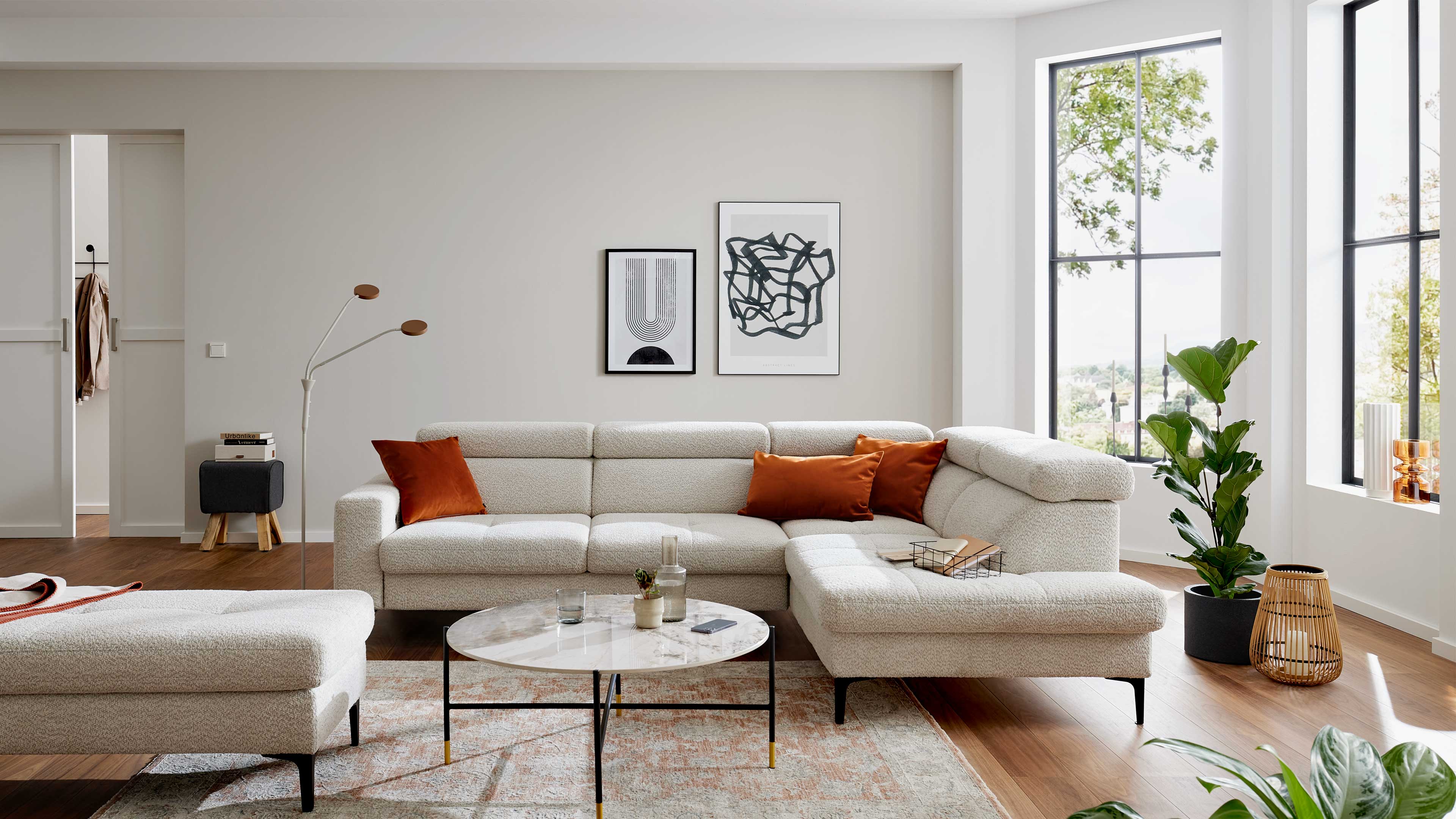 Ein großes cremefarbenes Ecksofa mit drei rostorangefarbenen Kissen steht vor einem runden Marmortisch, neben Fenster und Zimmerpflanzen.