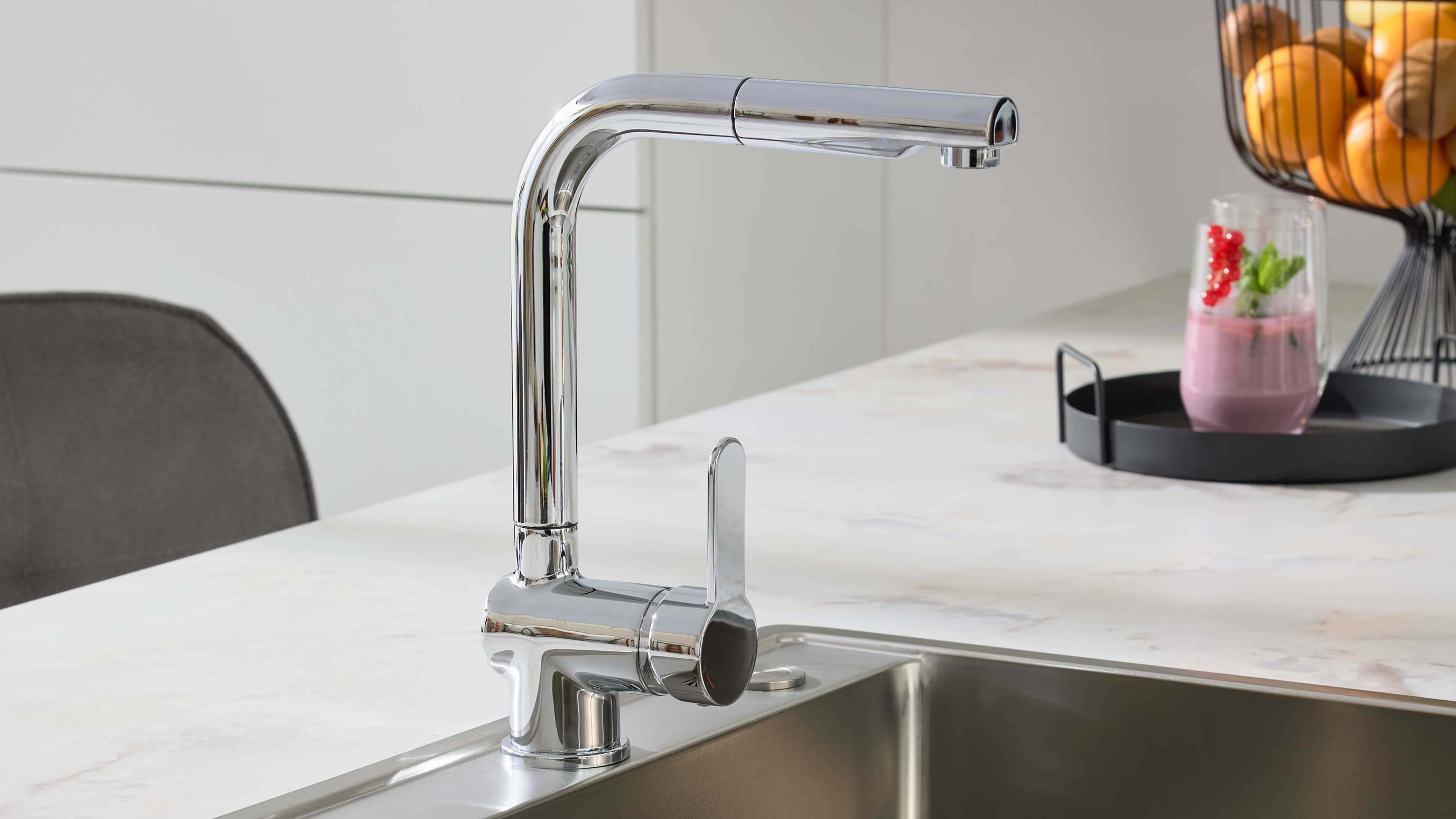 Verchromter Einhebel-Küchenmischer über eingebautem Edelstahlspülbecken auf heller Arbeitsplatte, rechts ein Tablett mit Glas und Obstkorb.