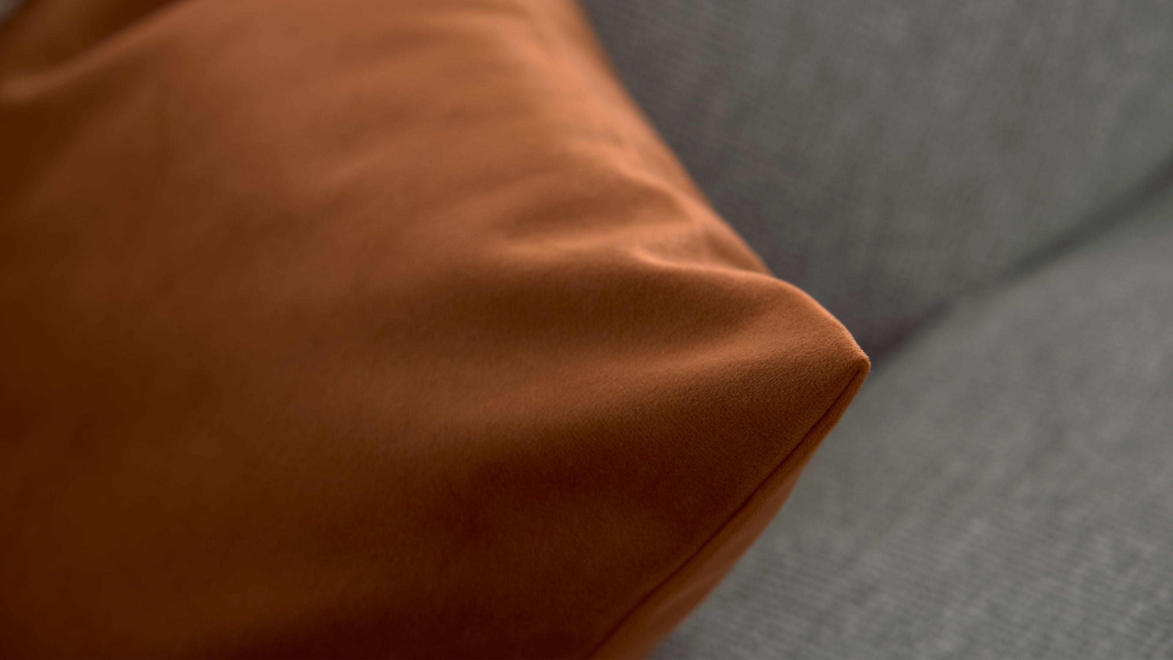 Rostbraunes Kissen mit glattem Stoff, Vorderkante scharf fokussiert, darauf ein hellgraues, fein gewebtes Sofa im Hintergrund.
