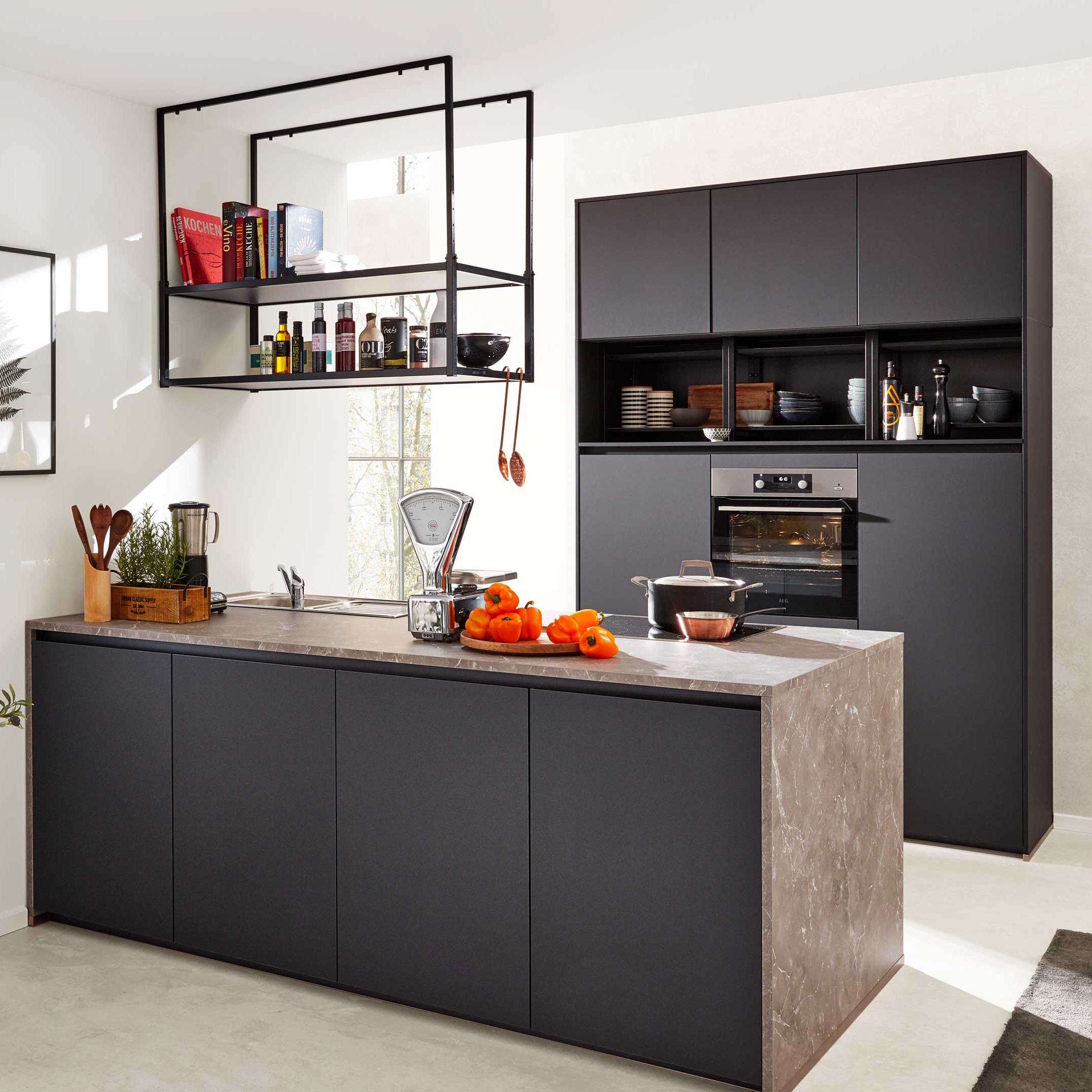 Schwarze Kücheninsel mit Marmorarbeitsplatte, hängendem Metallregal, Hochschrank mit Backofen, Waage und orangefarbenen Früchten.