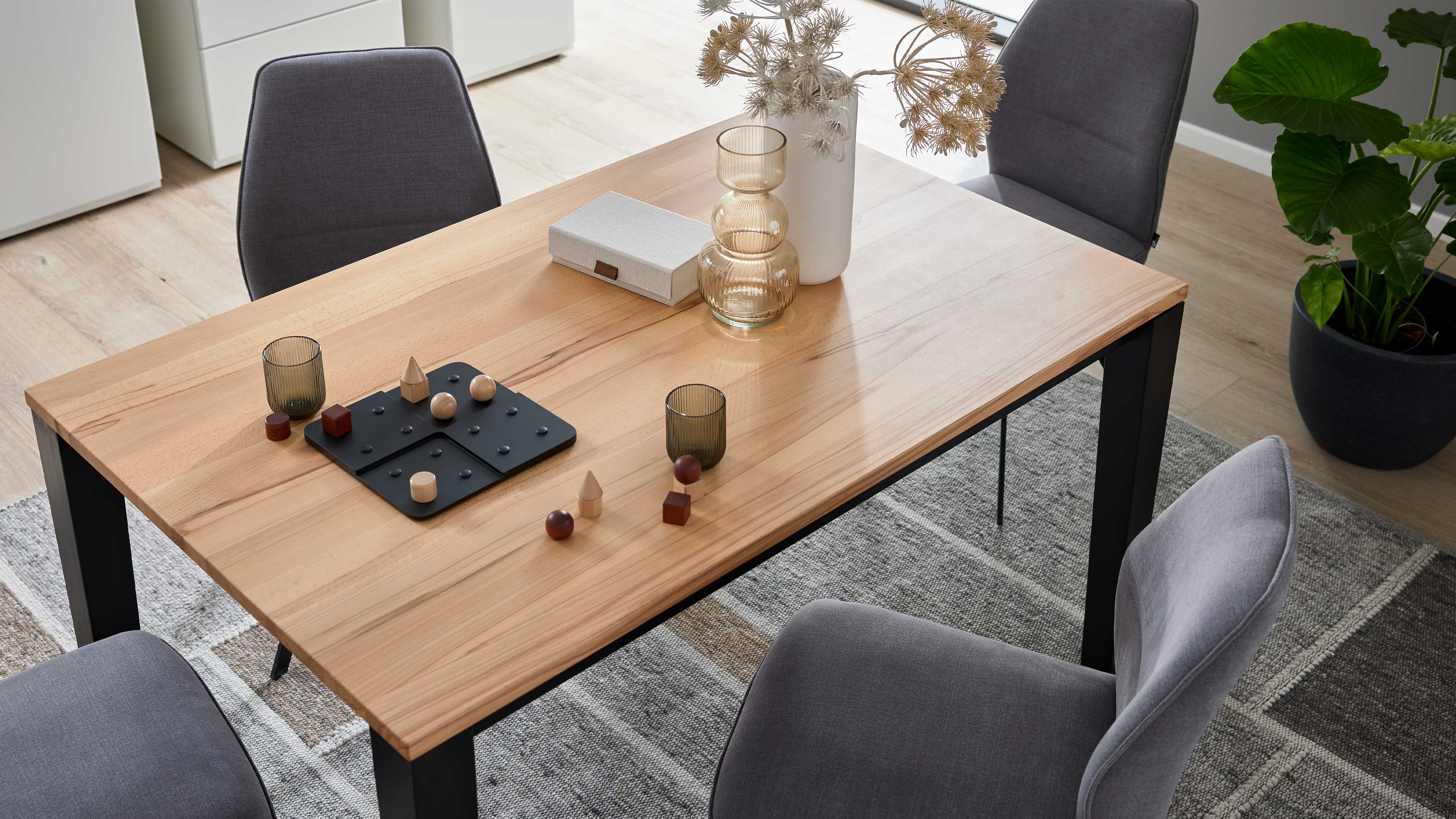 Holztisch mit schwarzen Metallbeinen, darauf Vase mit getrockneten Dolden, zwei Gläser, Spielbrett mit Holzfiguren und graue Stühle.