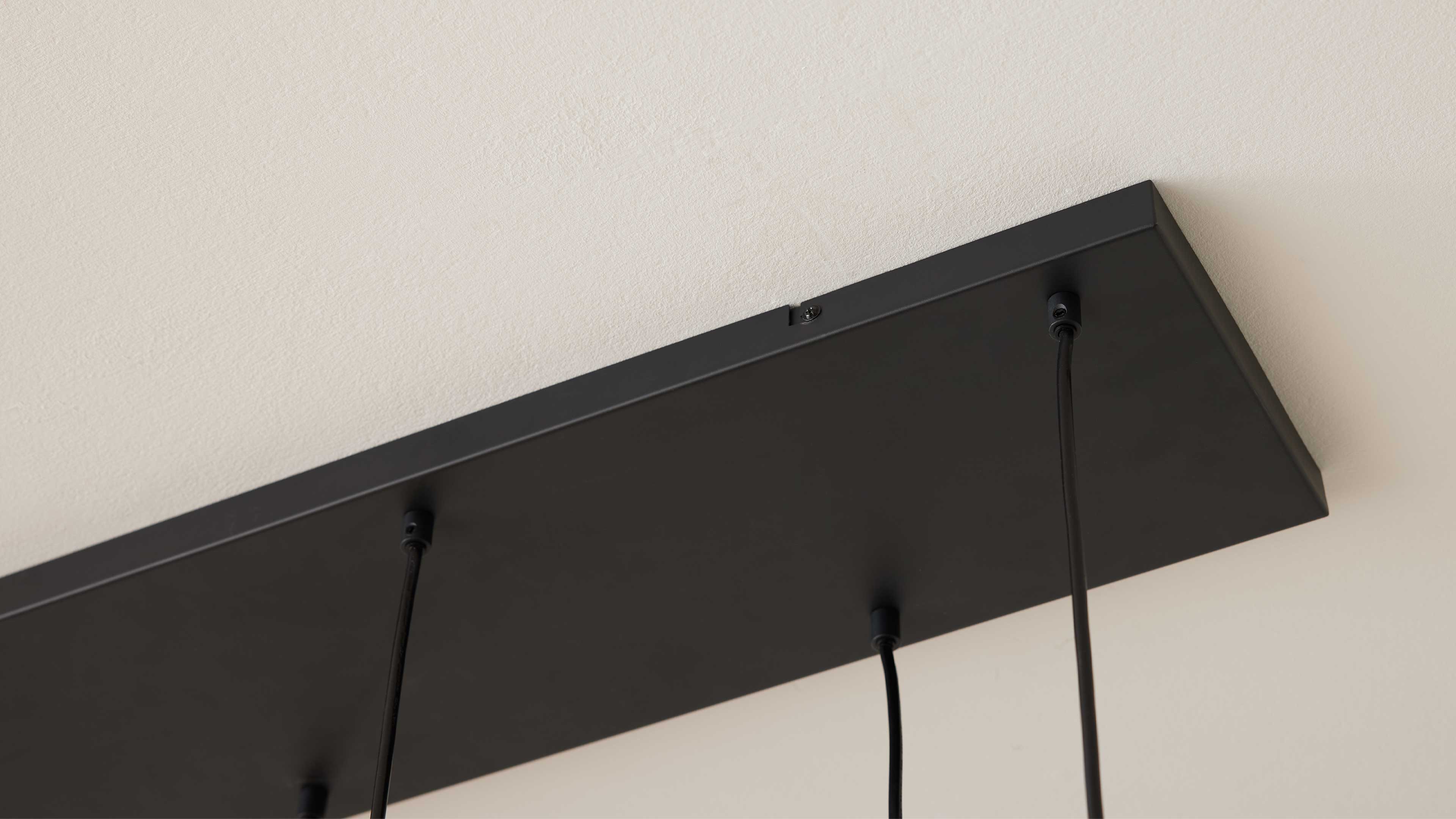 Schwarze rechteckige Deckenhalterung mit drei vertikal herabhängenden schwarzen Kabeln an weißer, leicht strukturierter Decke.