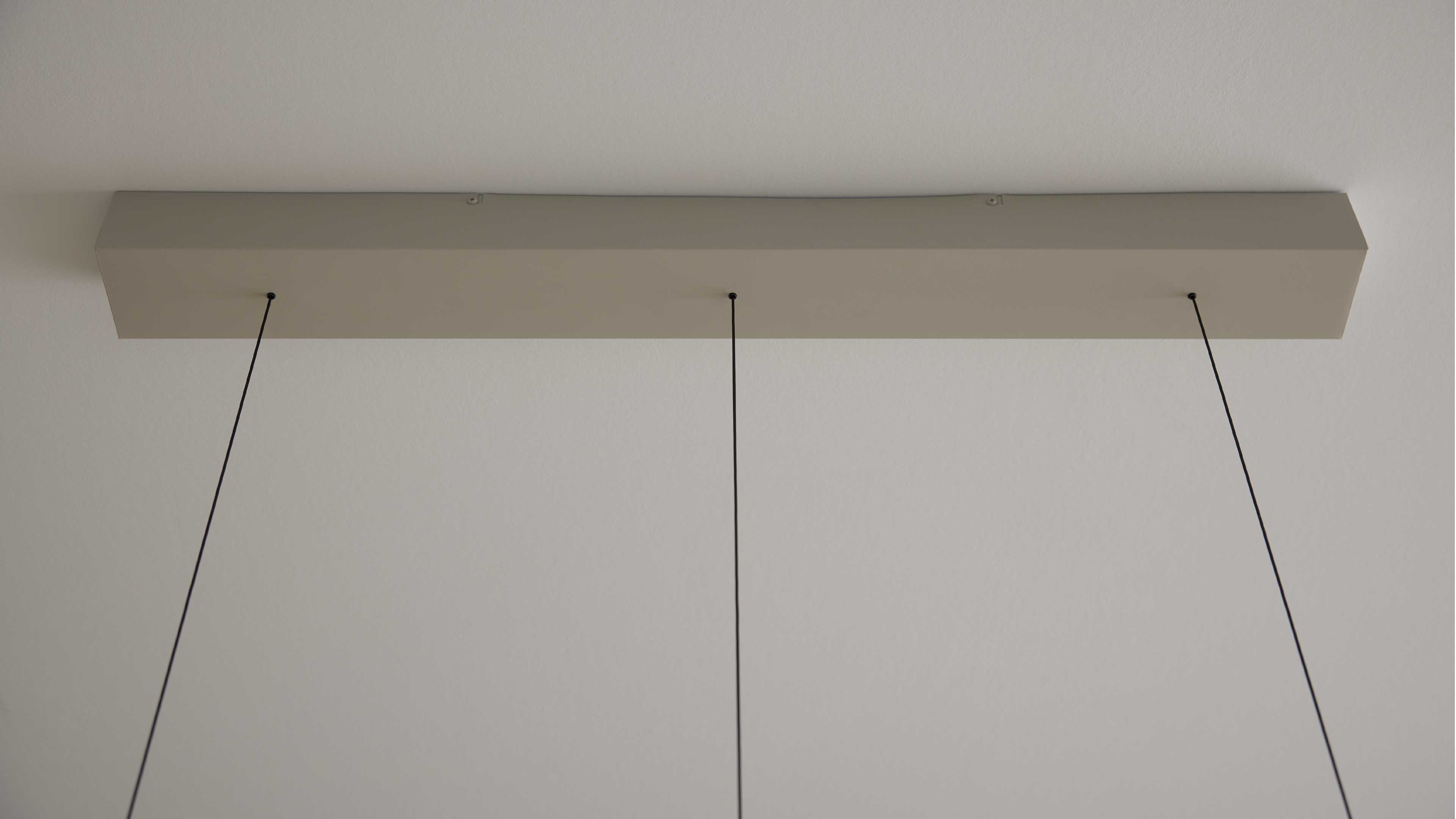 Rechteckiges hellbeiges Deckenpaneel an weißer Decke, daran drei dünne schwarze Kabel in gleichen Abständen hängend.