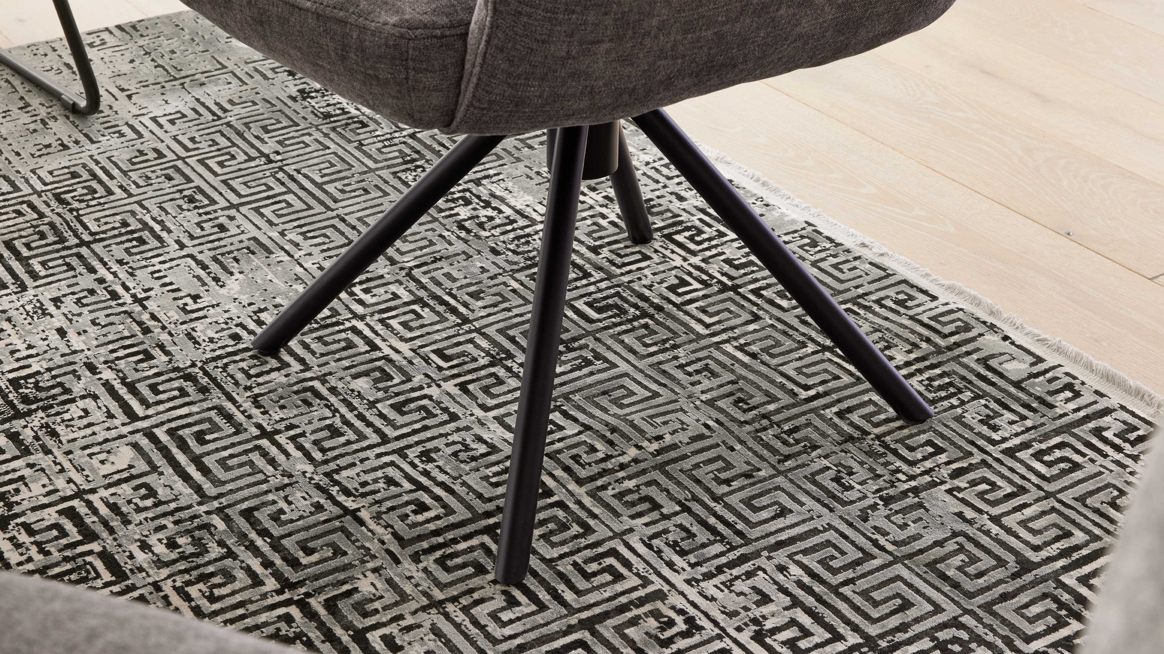 Grau gepolsterter Drehstuhl mit schwarzen Metallbeinen steht auf einem schwarz-grauen Teppich mit geometrischem Muster und Fransen.