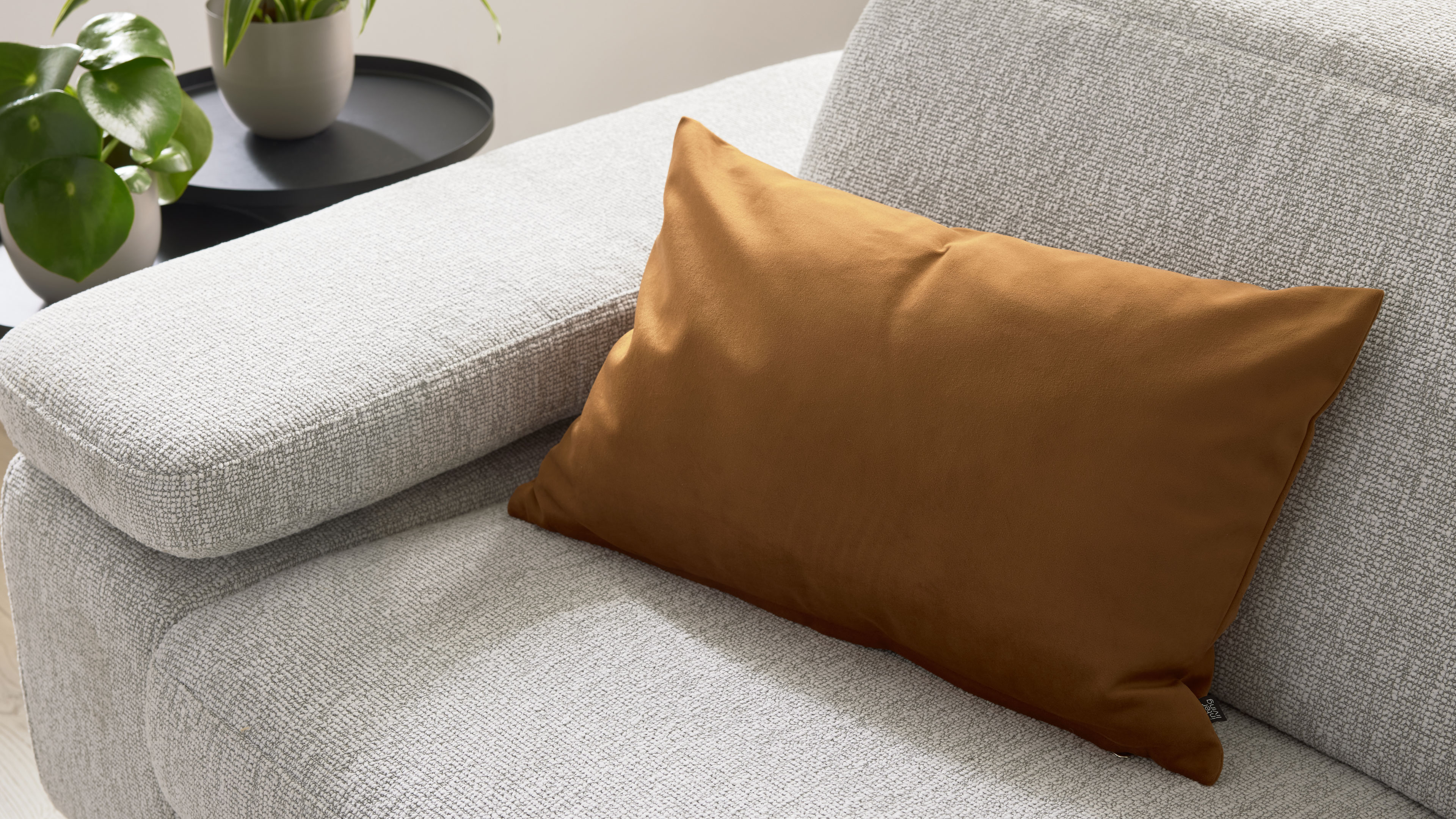 Hellgraues, strukturiertes Sofa mit breiter Armlehne und einem rechteckigen braunen Kissen, seitlich zwei Pflanzen.