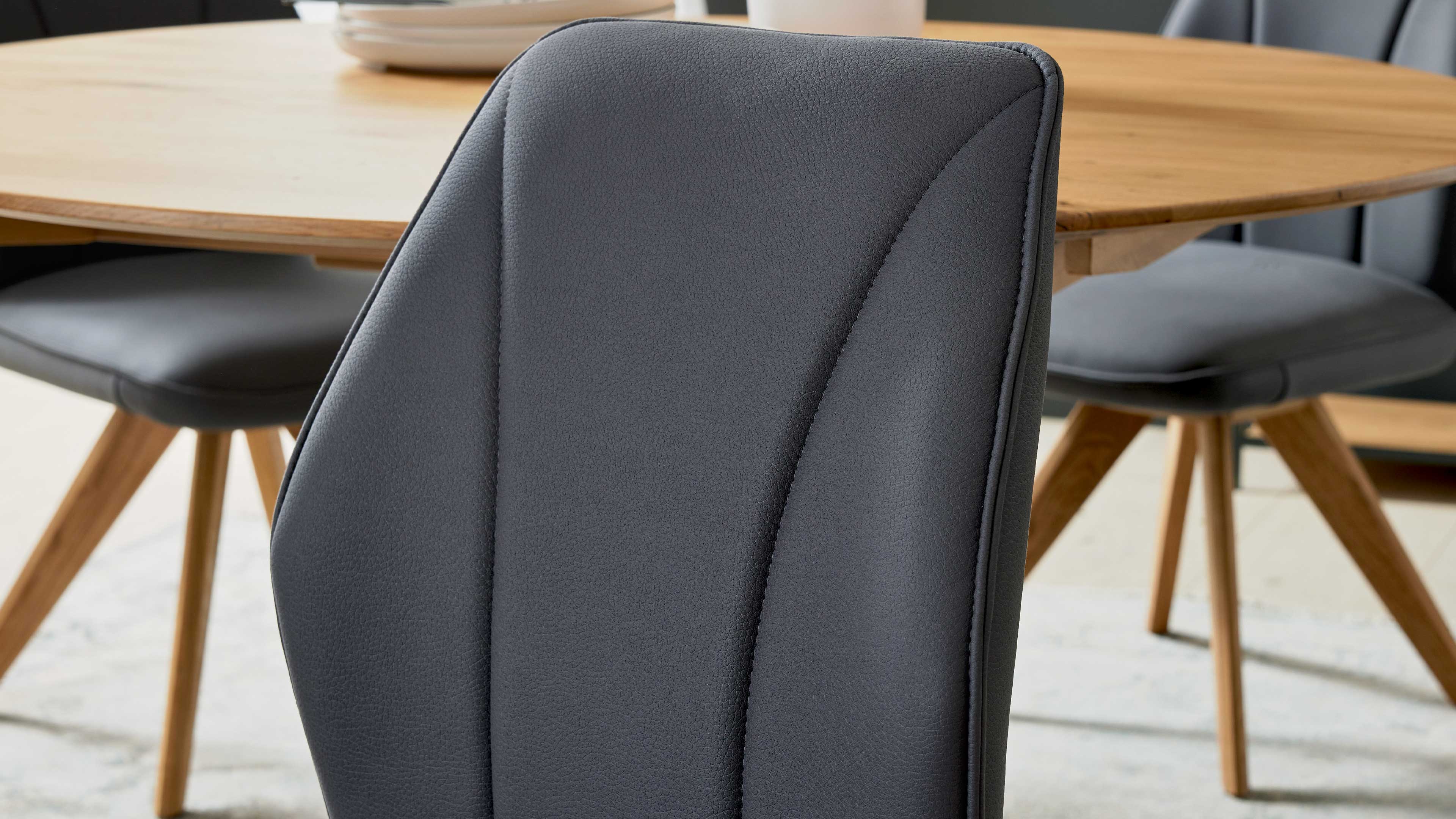 Nahaufnahme der dunkelgrauen, gesteppte Rückenlehne eines Stuhls mit senkrechten Nähten, im Hintergrund runder Holztisch und Holzbeine.