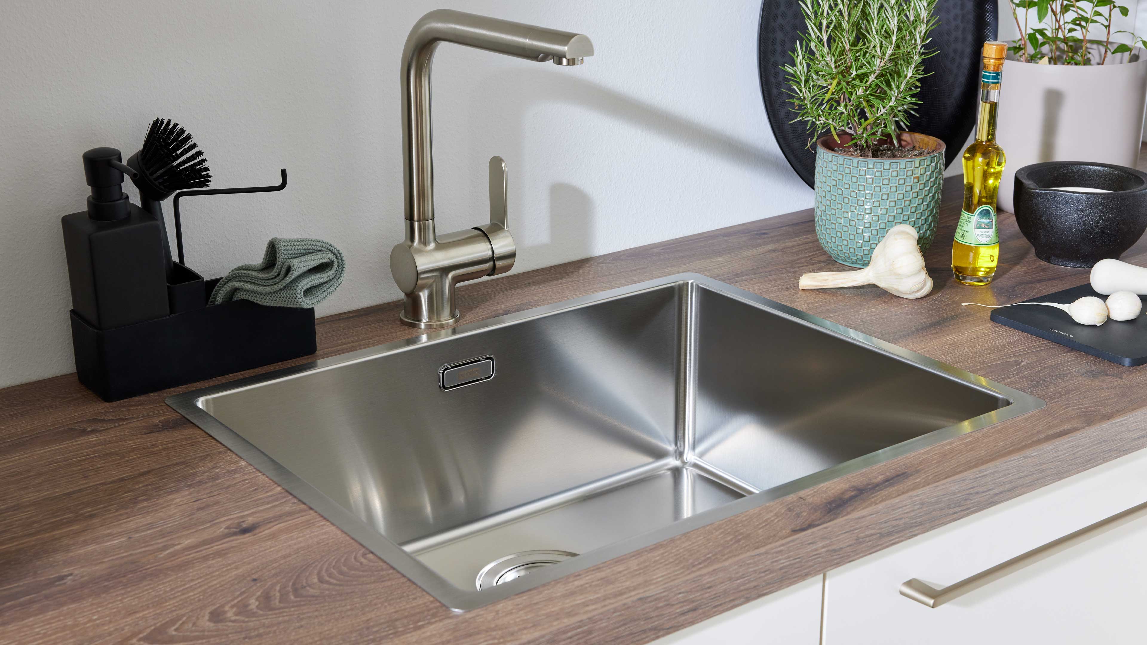 Edelstahl-Spüle mit hohem Wasserhahn in Holzarbeitsplatte, links Seifenspender mit Bürste, rechts Topf mit Pflanze, Ölflasche und Knoblauch.
