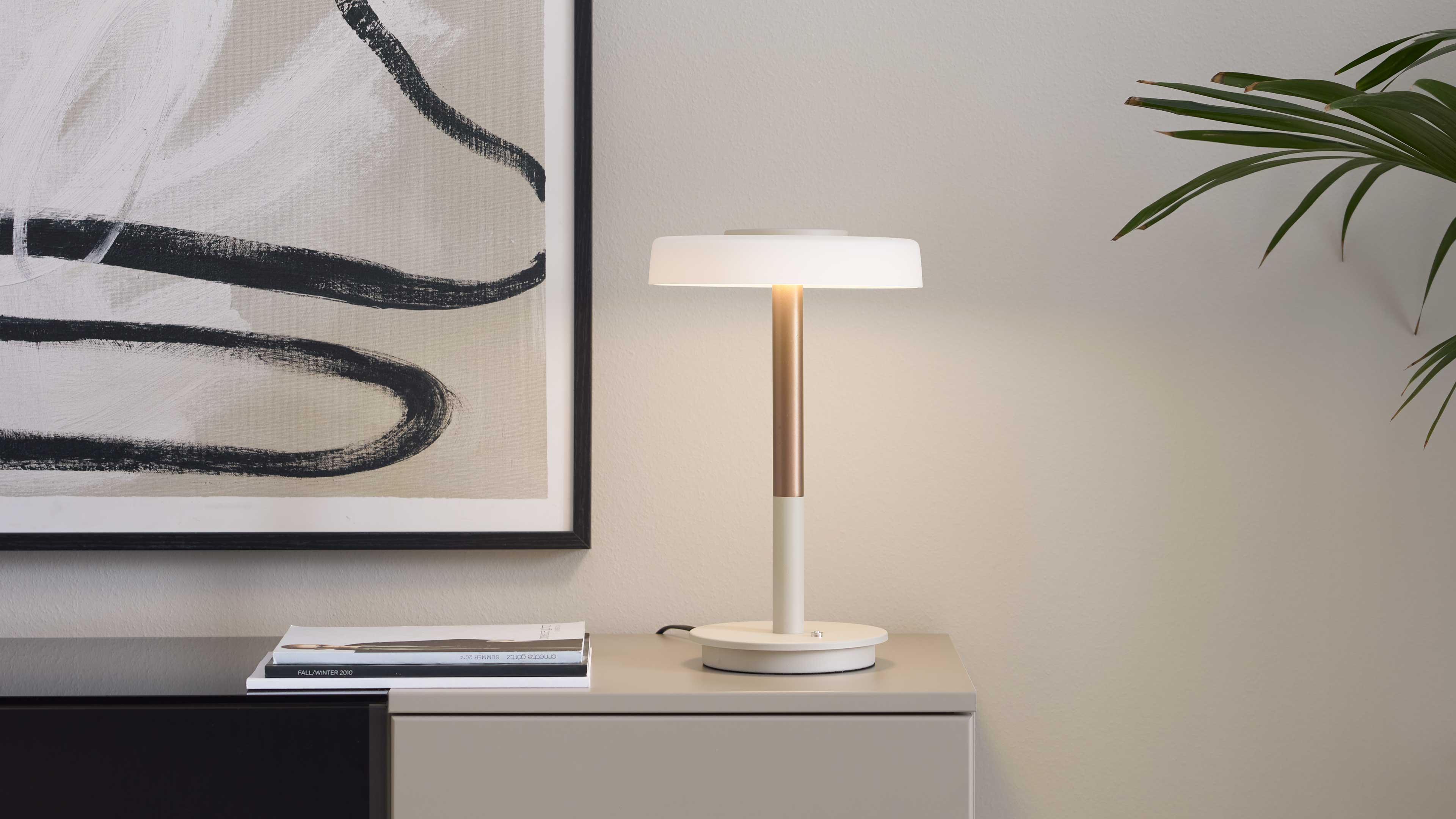 Tischlampe mit rundem weißem Schirm, goldfarbenem Schaft und Basis auf Sideboard neben gerahmtem abstrakten Bild, Heften und Pflanzenblatt.