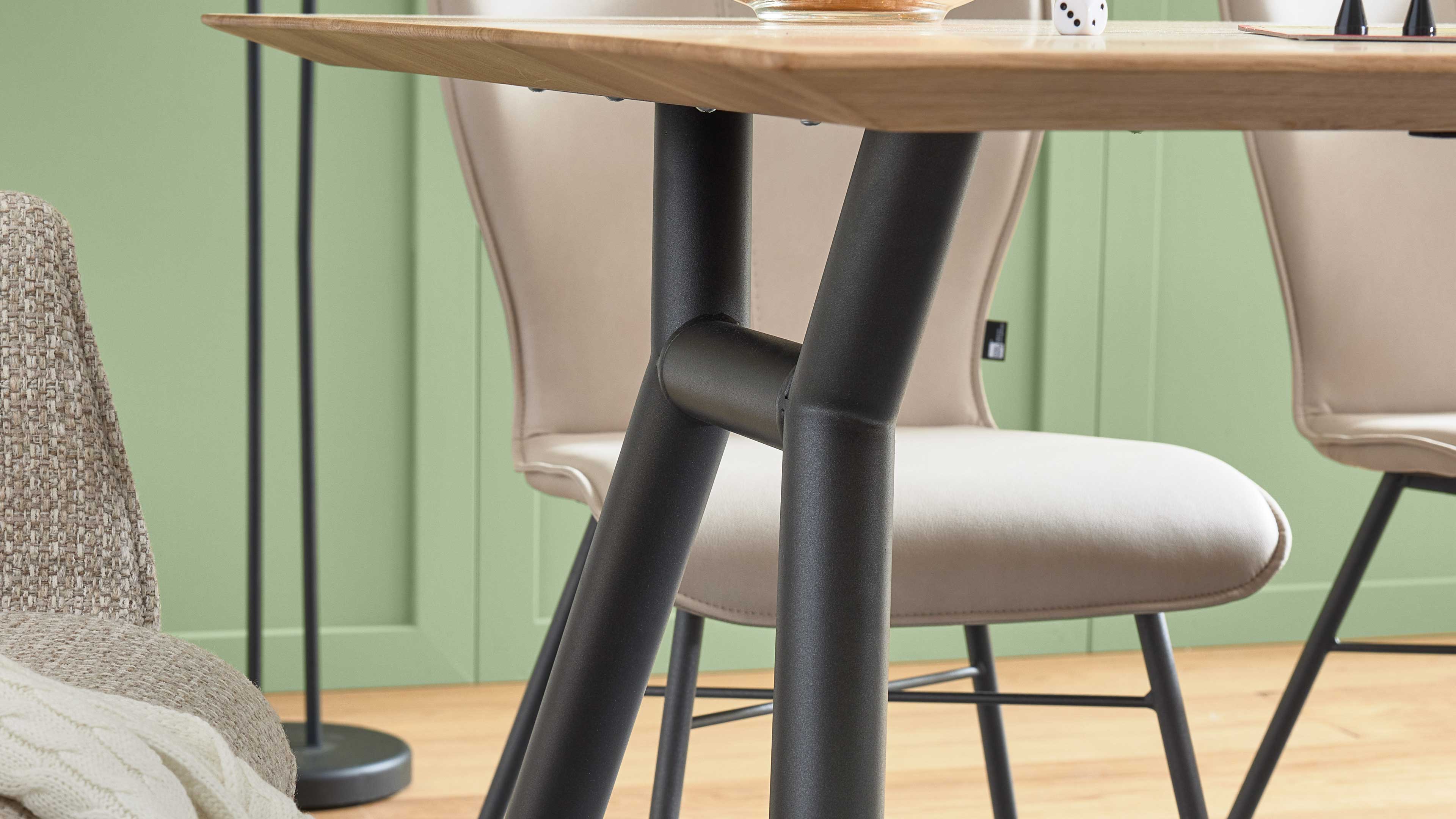 Schwarzes, gekreuztes Metall-Gestell trägt eine hellholz-Platte, im Hintergrund beige gepolsterte Stühle vor grüner Wand.