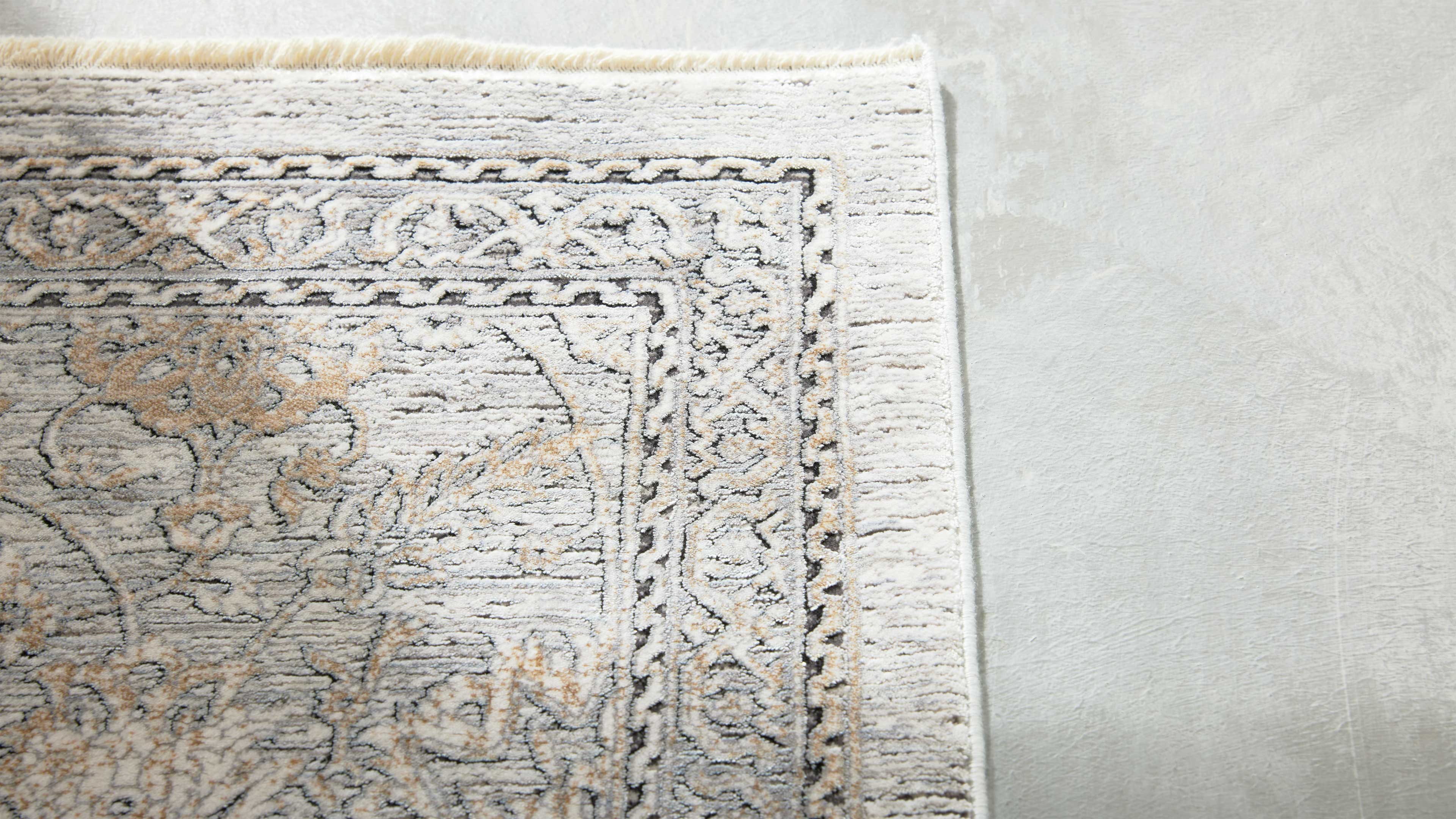 Ecke eines hellen Teppichs mit Fransenrand, ornamentaler Bordüre und floralen Mustern in Beige und Grau auf hellem Boden.