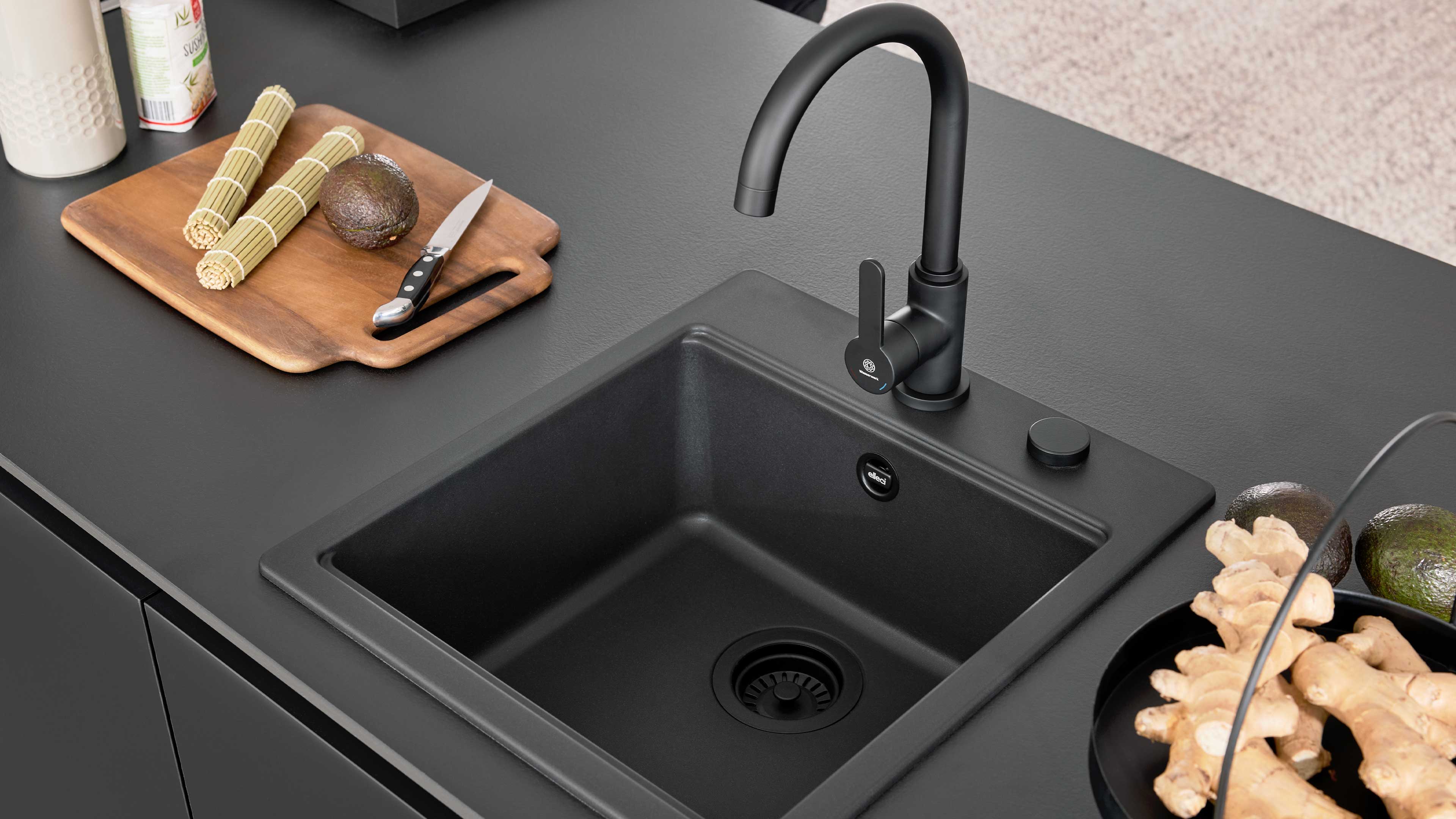 Schwarzes Einbauspülbecken mit gebogener Armatur auf mattschwarzer Arbeitsplatte, links Holzbrett mit Messer und Avocado, rechts Ingwer.