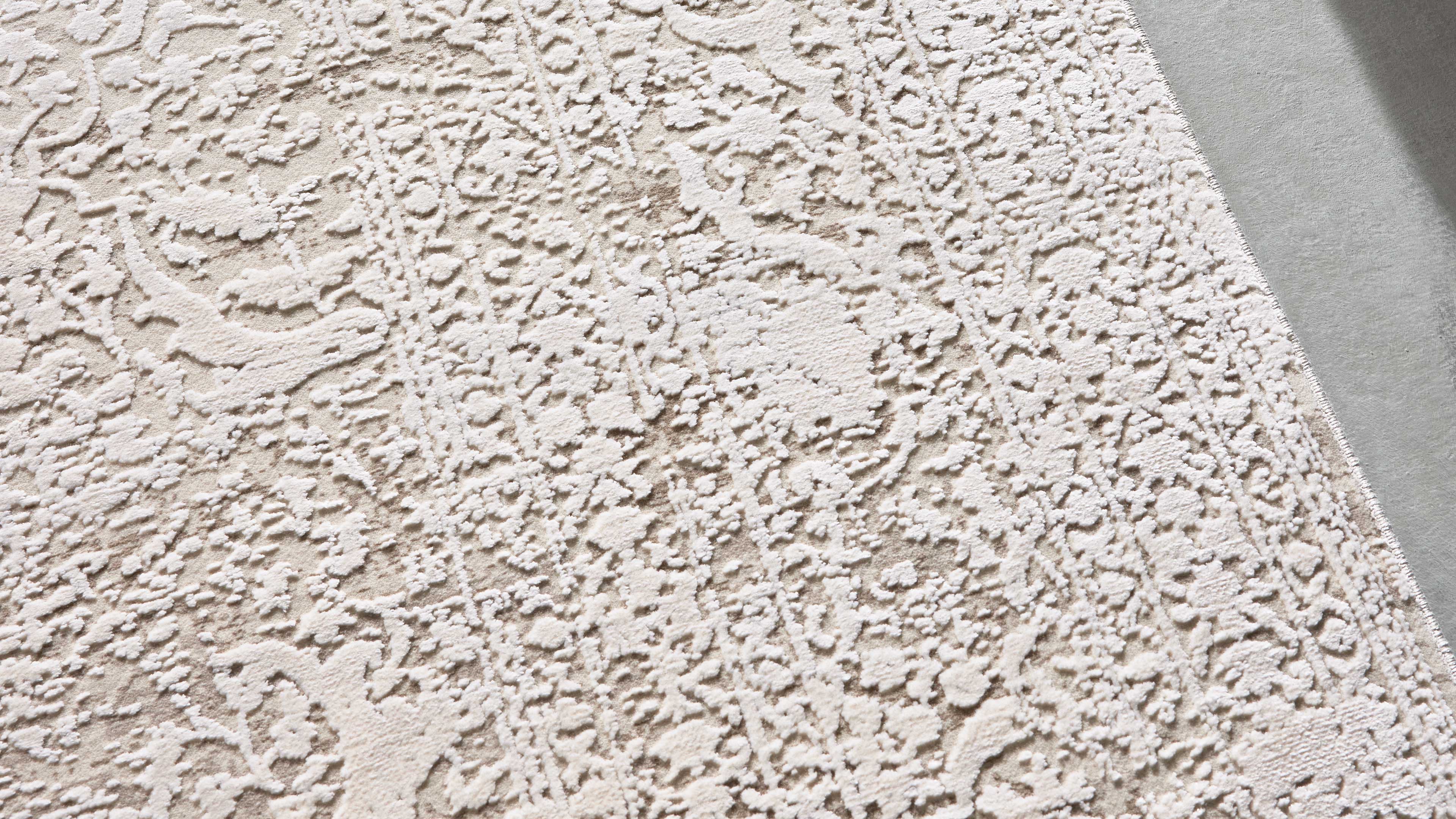 Heller Teppich mit reliefartigem, floralen Ornamentmuster in Creme- und Beigetönen, rechts sichtbare Saumnaht und Boden.