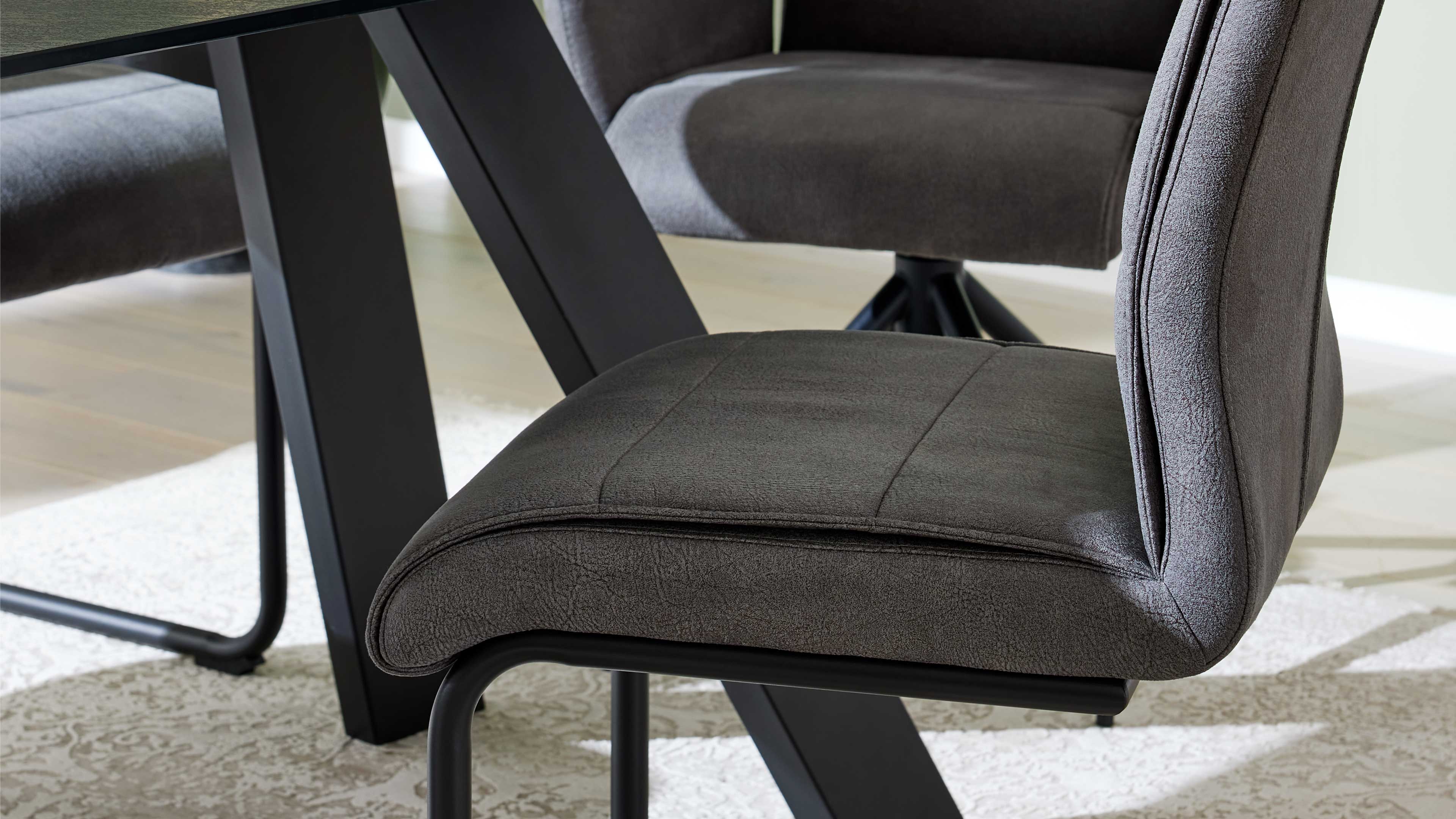 Gepolsterter dunkelgrauer Stuhl mit konturierter Sitzfläche und geschwungener Rückenlehne auf schwarzem Metallgestell, neben Tischbein.