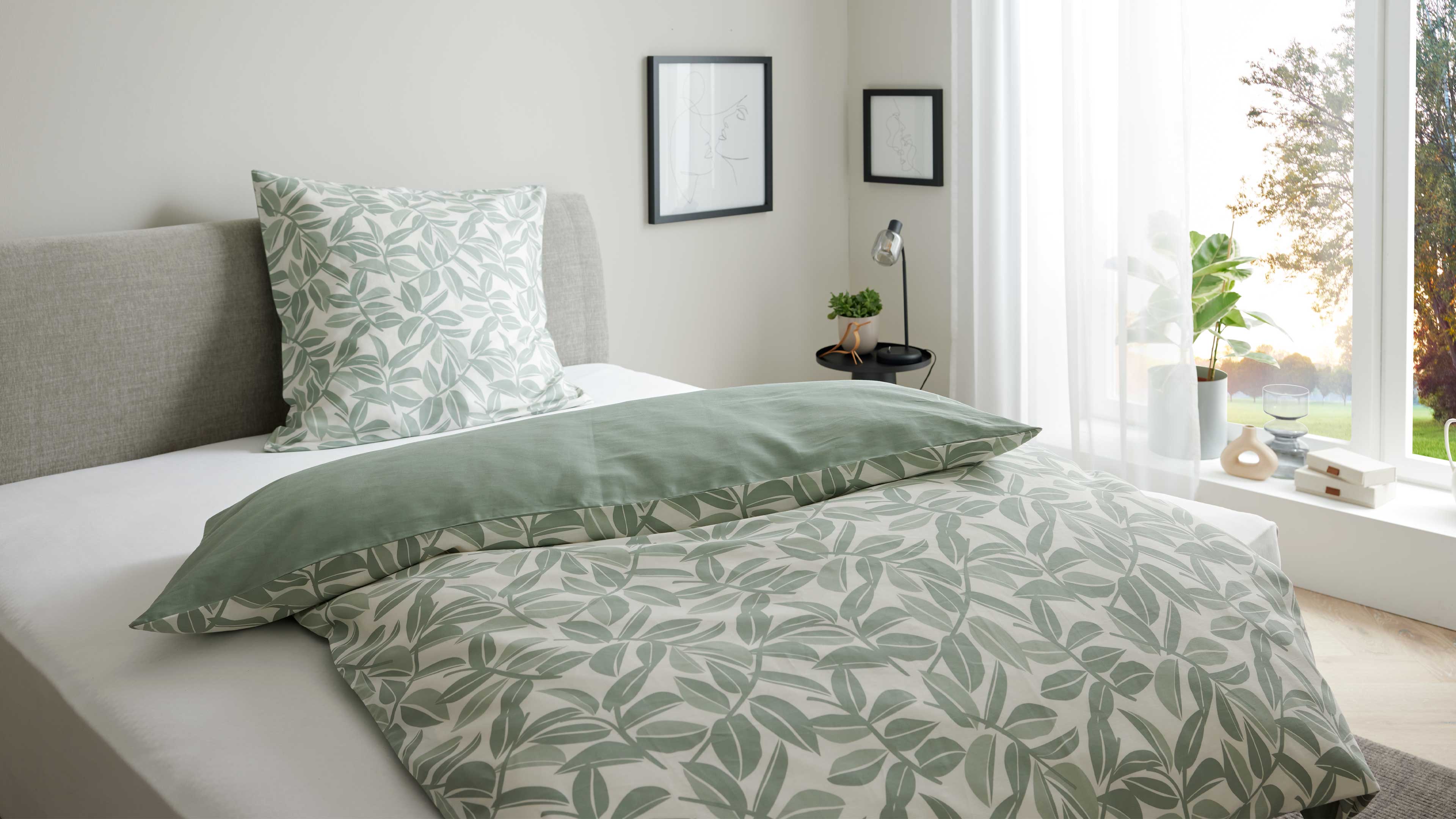 Bett mit grauem Polsterkopfteil, grün-weiß gemustertem Blatt-Bettbezug und Kissen, kleiner Nachttisch mit Lampe und Pflanze.